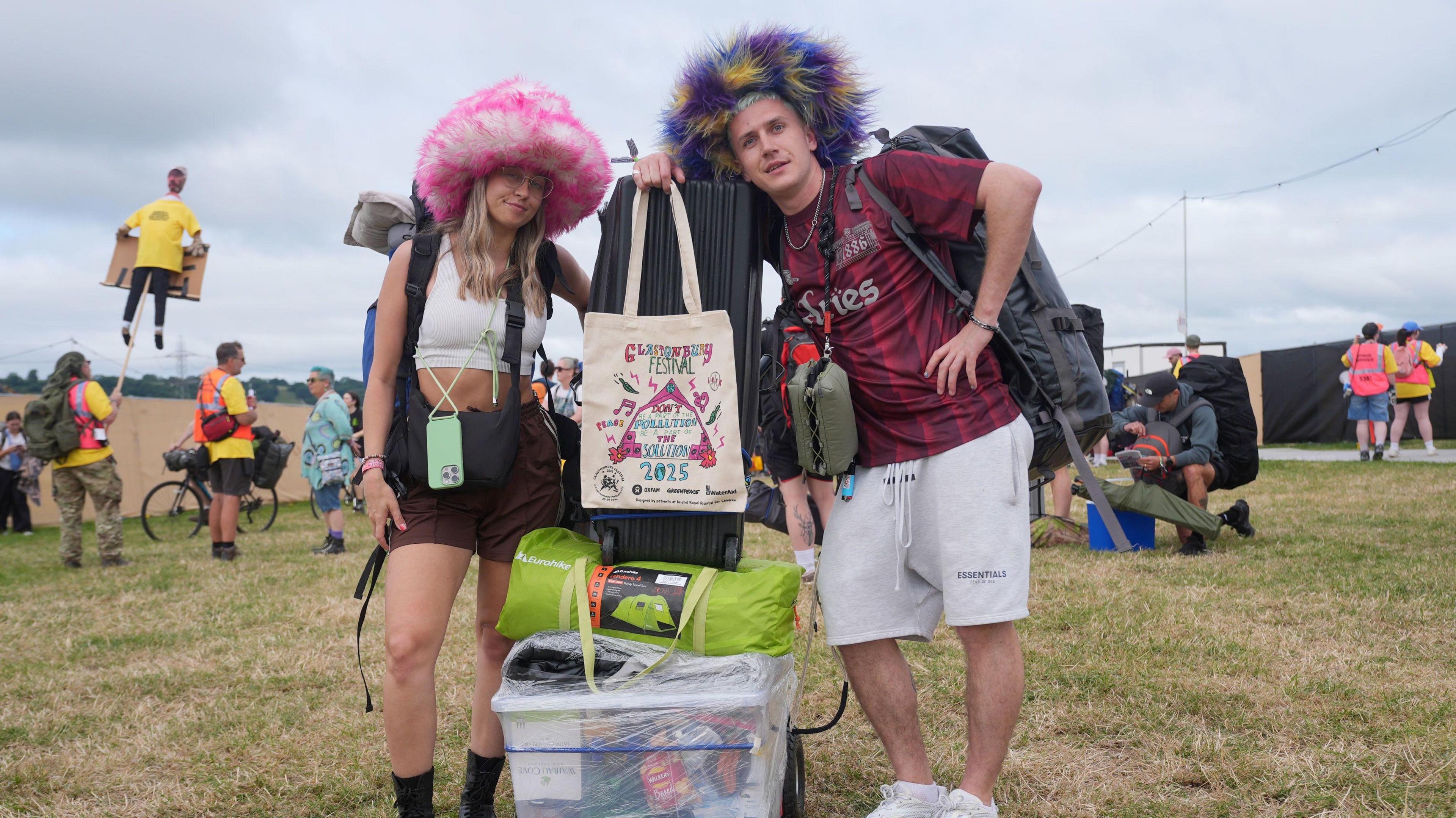 Two people pose with a trolley and their suitcase carrying all their festival equipment into the site. They are both wearing huge furry hats. They are posing with a white Glastonbury 2025 tote bag. Festival staff and volunteers can be seen in the distance wearing high vis jackets.