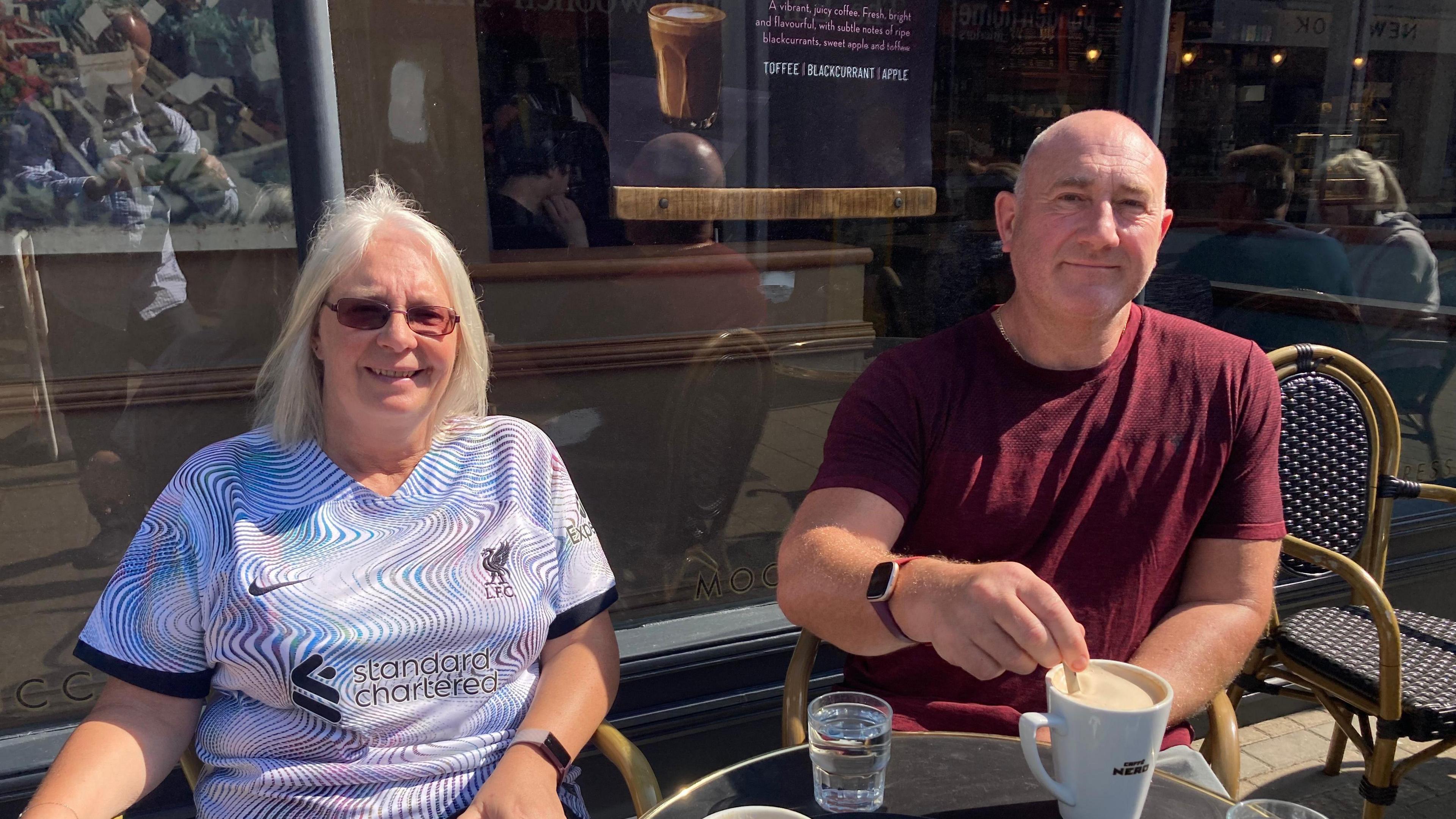 Susan and Wayne Thirwell sit at an outdoor table at a coffee shop both smiling at the camera