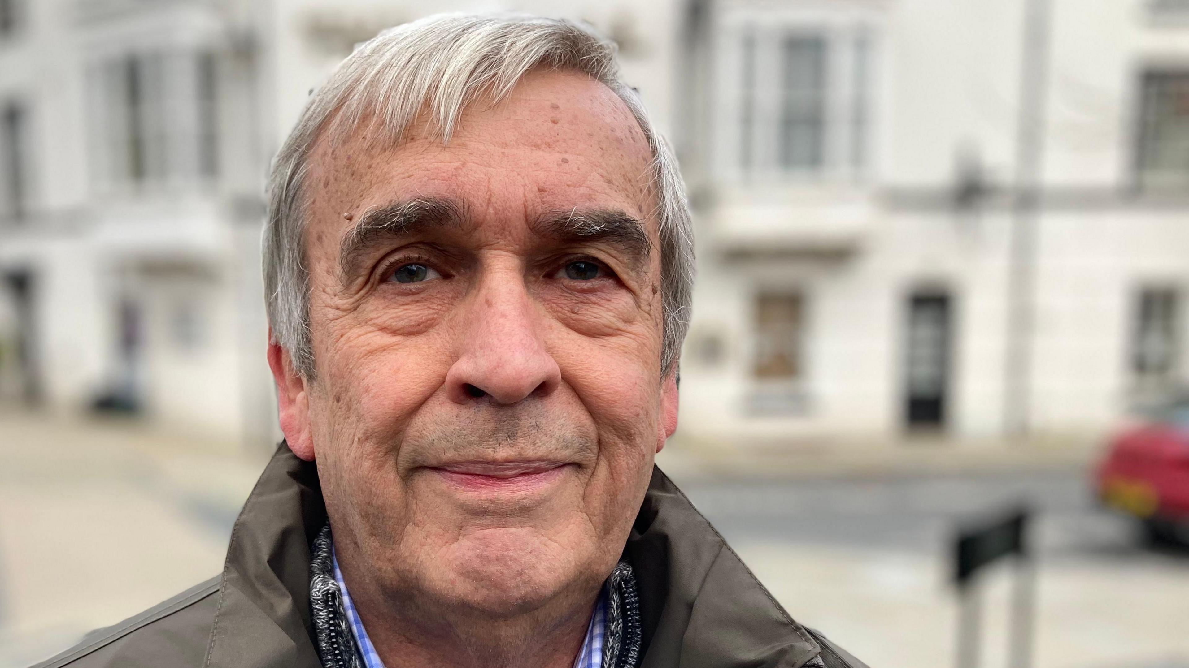 Portrait of Martyn Holland, white and grey haired man wearing a dark green coat and blue checked shirt, staring directly into the shot.