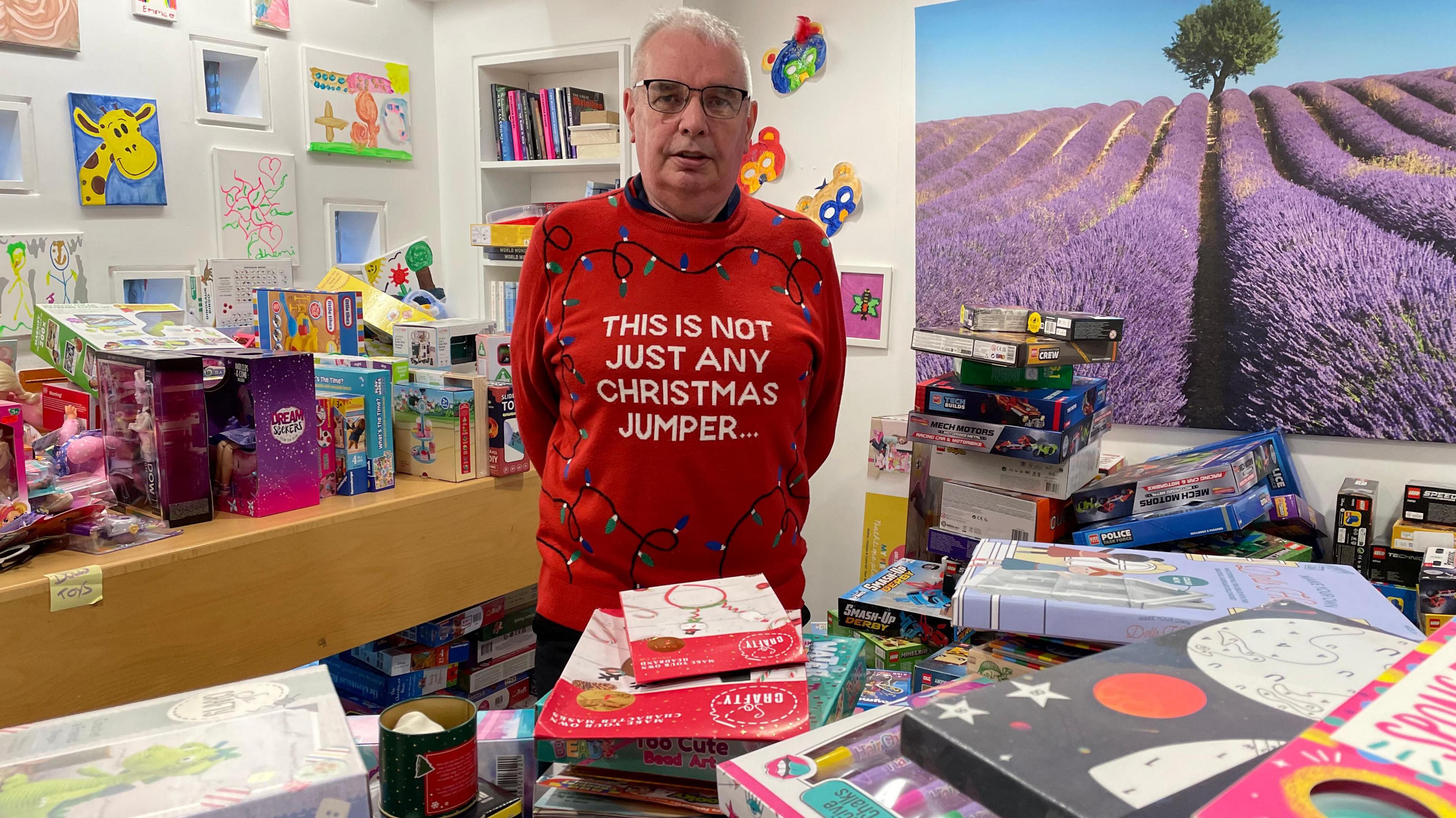 Colin O'Flaherty standing in a room full of children's toys, books and games. He is wearing a red jumper with Christmas lights on it and the words This is not just any Christmas jumper. He has grey hair and glasses and is looking at the camera. There is a large picture on the wall behind him showing a purple flowers and a tree in the distance.