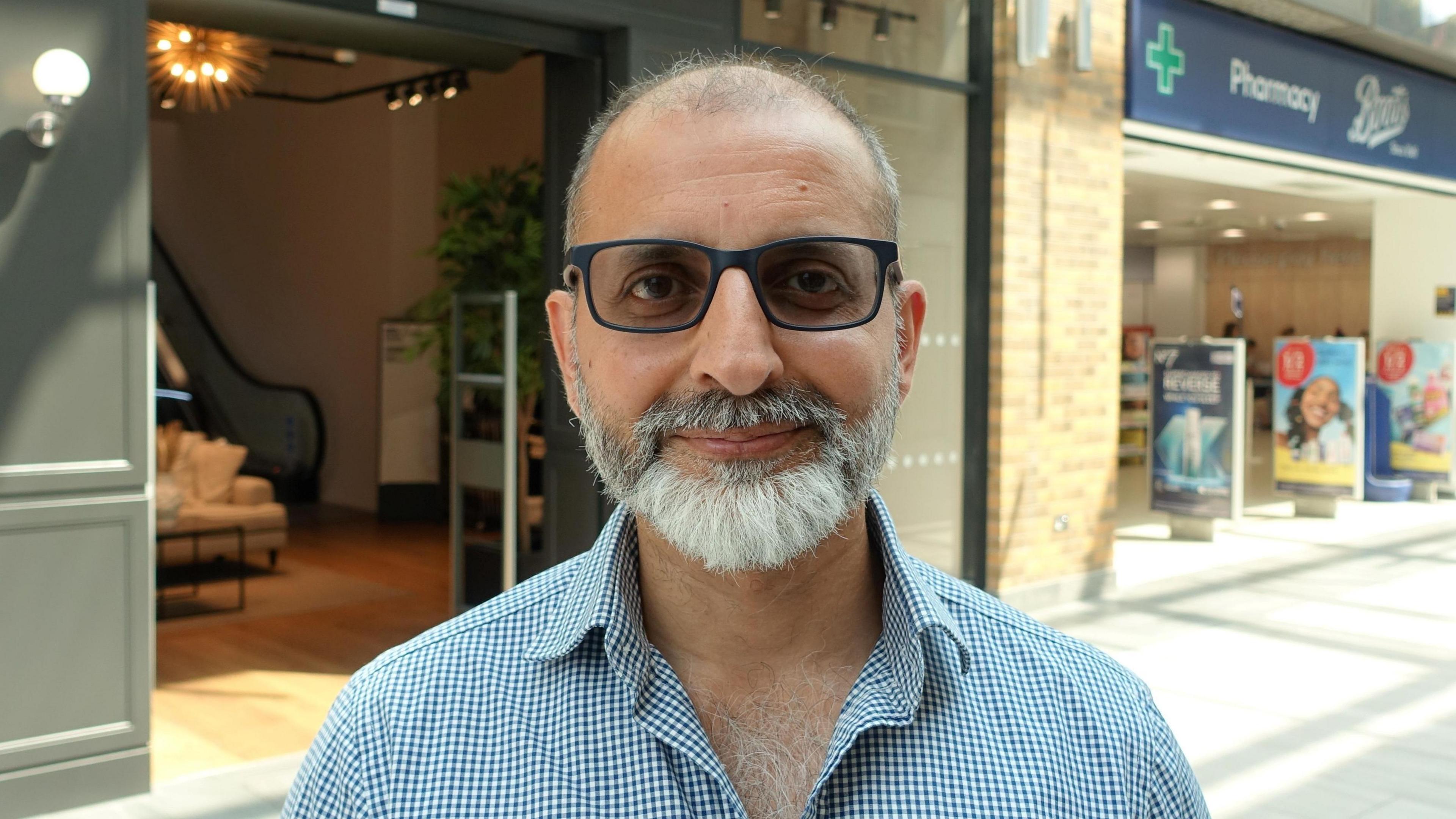 Head-and-shoulders photo of Subjit Jassy wearing a blue and white shirt and glasses, in the background are Grafton Centre shops.
