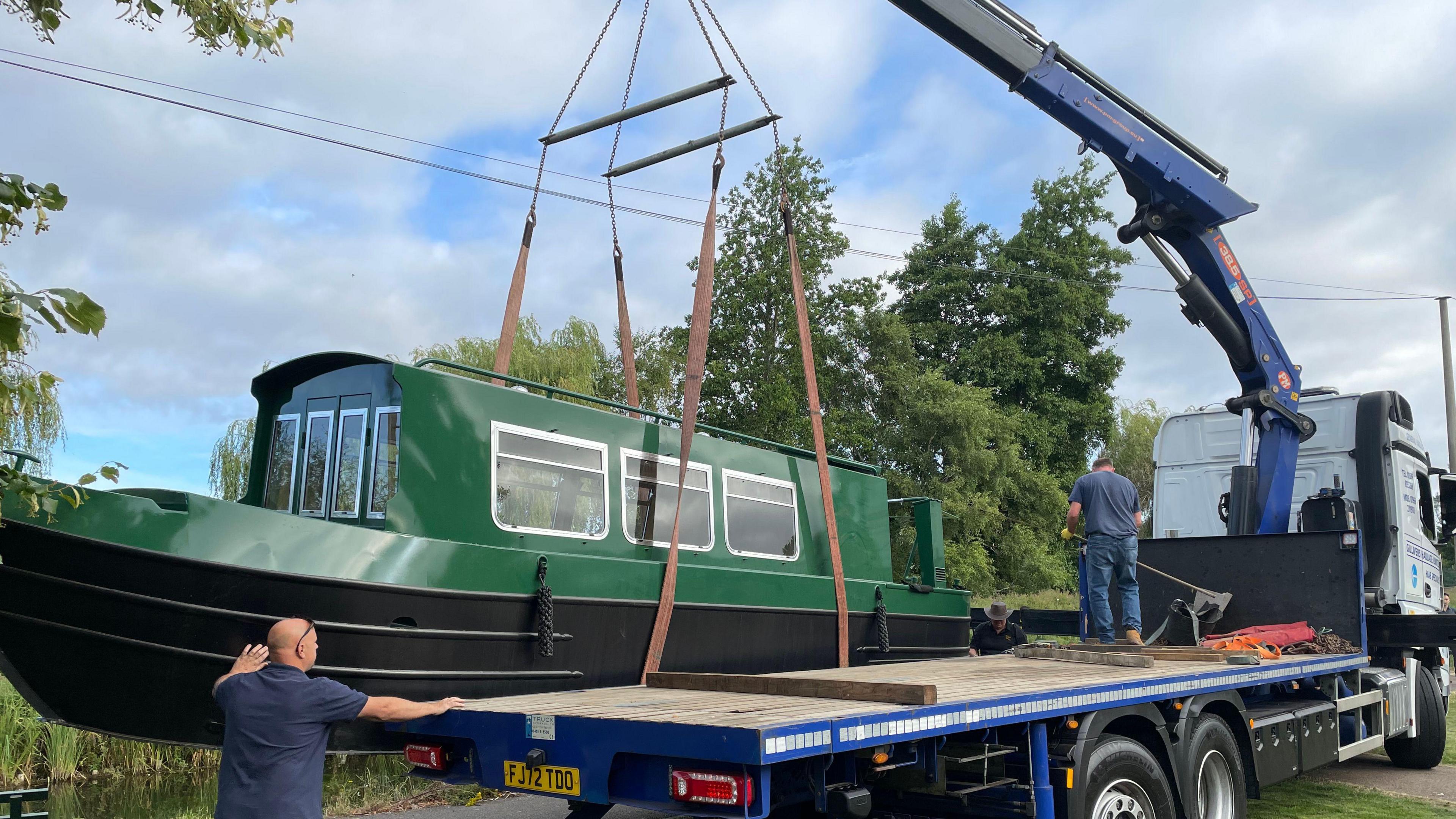 A green narrowboat hoisted on red straps is lowered into the river by a blue crane.