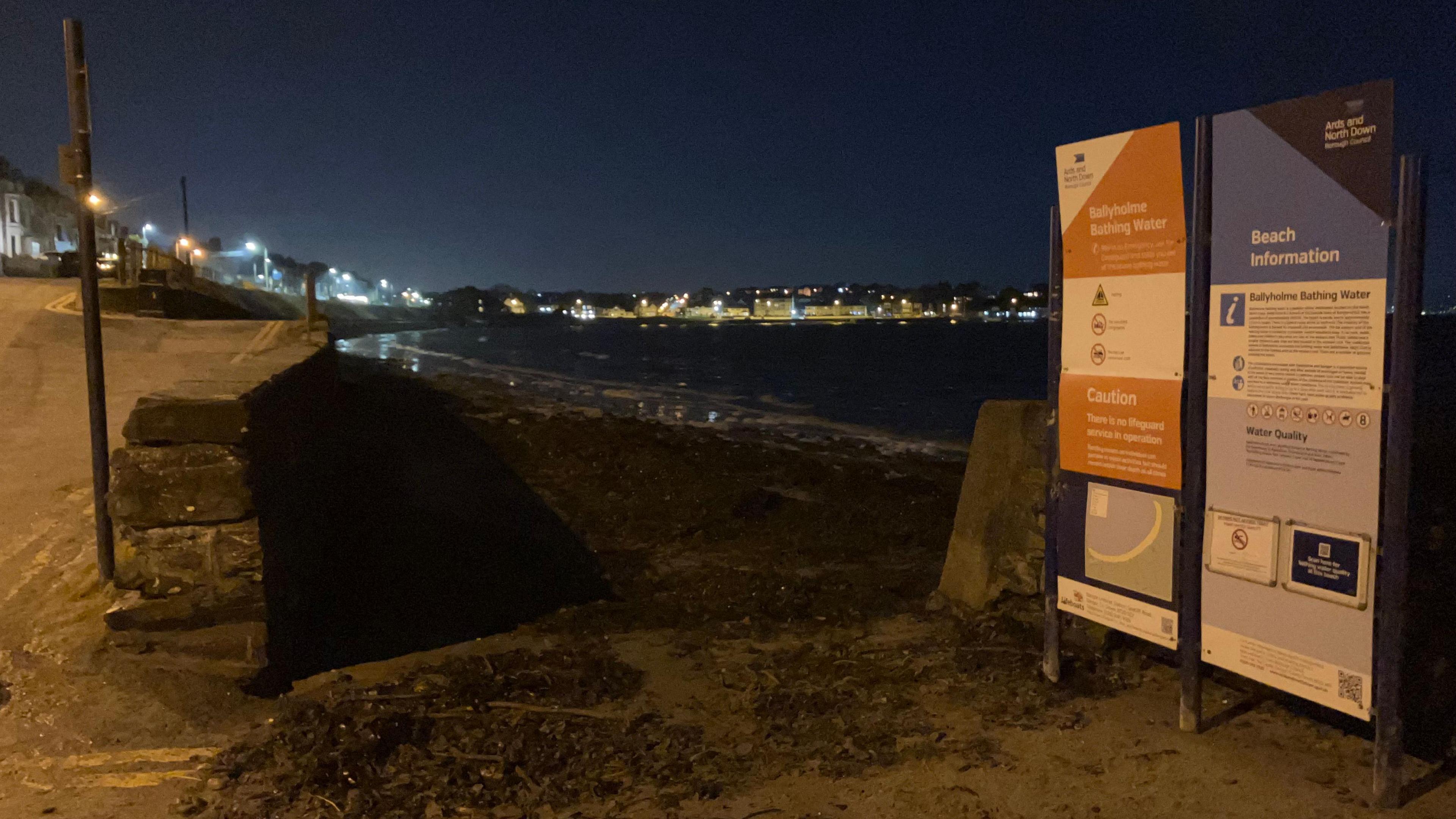 A beach at night time. In the background a headland with the lights of houses. A stone wall at the top of the beach and on the other wise a road. Signs at the entrance to the beach read "ballyholme bathing water" and "beach information" 