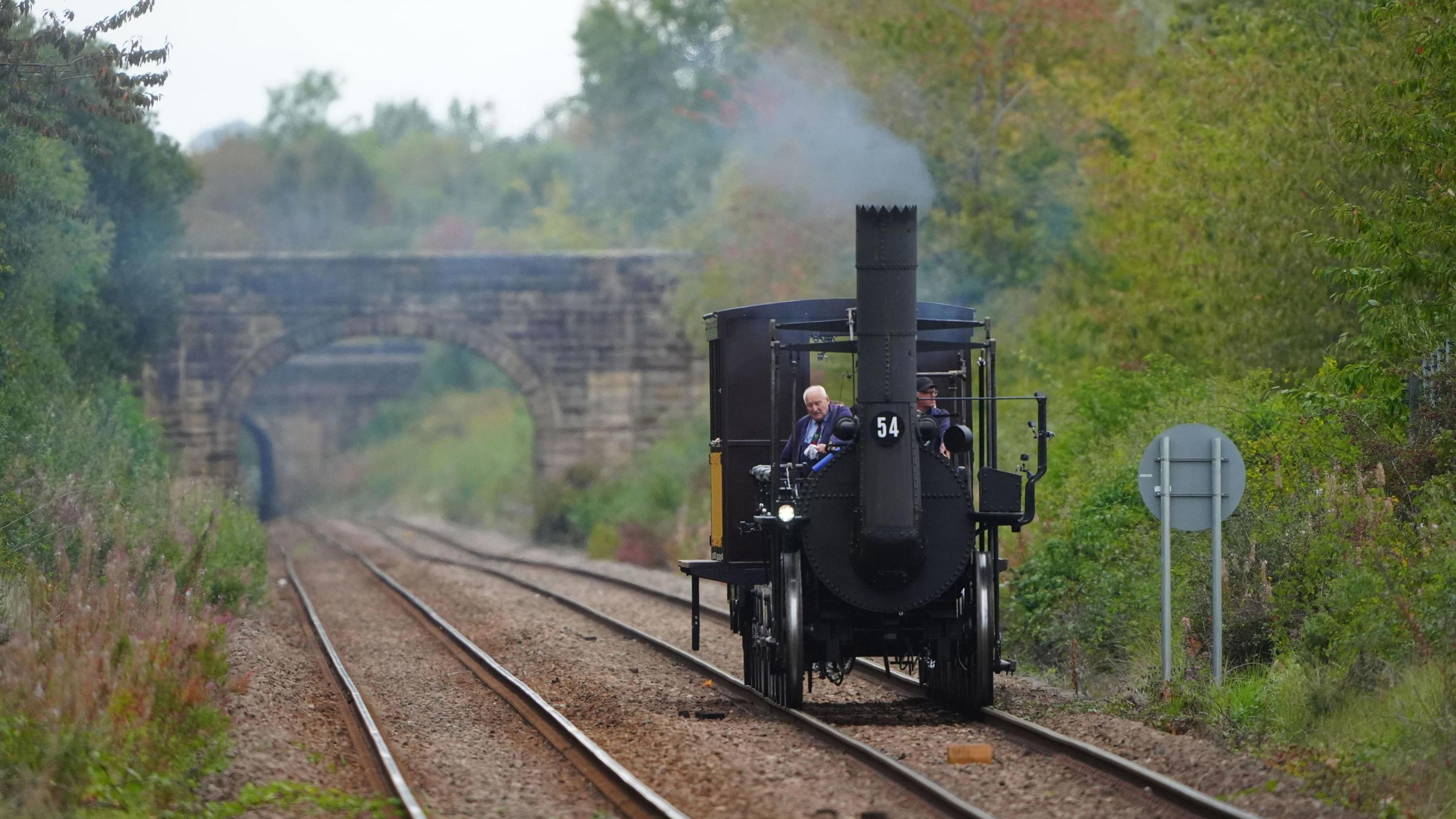 Replica Locomotion No1 approaches Heighington station, County Durham, on its first official journey in 200 years from the Locomotion Museum in Shildon to Darlington, as part of celebrations marking the 200th anniversary of British passenger rail services. Two men are on board. The track is empty and surrounded by overgrown shrubbery. The black train has a funnel at the front with steam coming out.