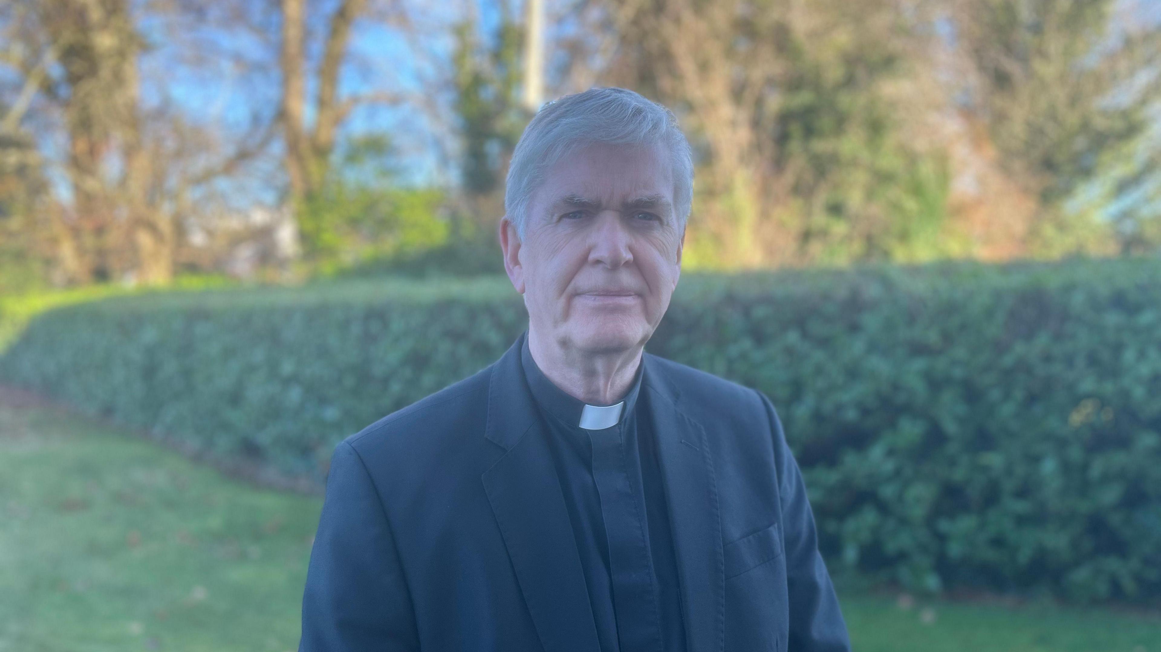 A priest is looking at the camera. He's standing on grass. He has grey hair. He has a neutral expression. He's wearing his black priest suit with a white collar. There are bushes and trees behind him. 