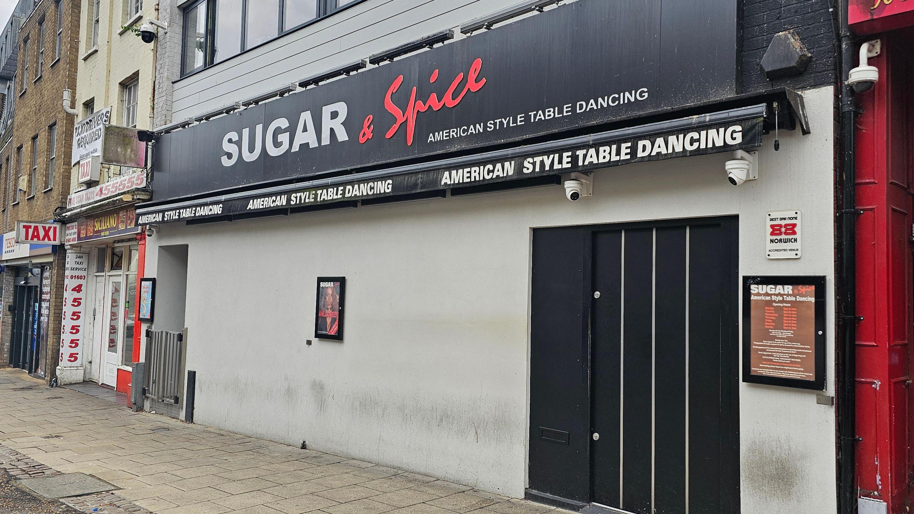 The outside of Sugar and Spice. A grey building with a black door and black sign which has grey and red writing. It says it has American style table dancing.