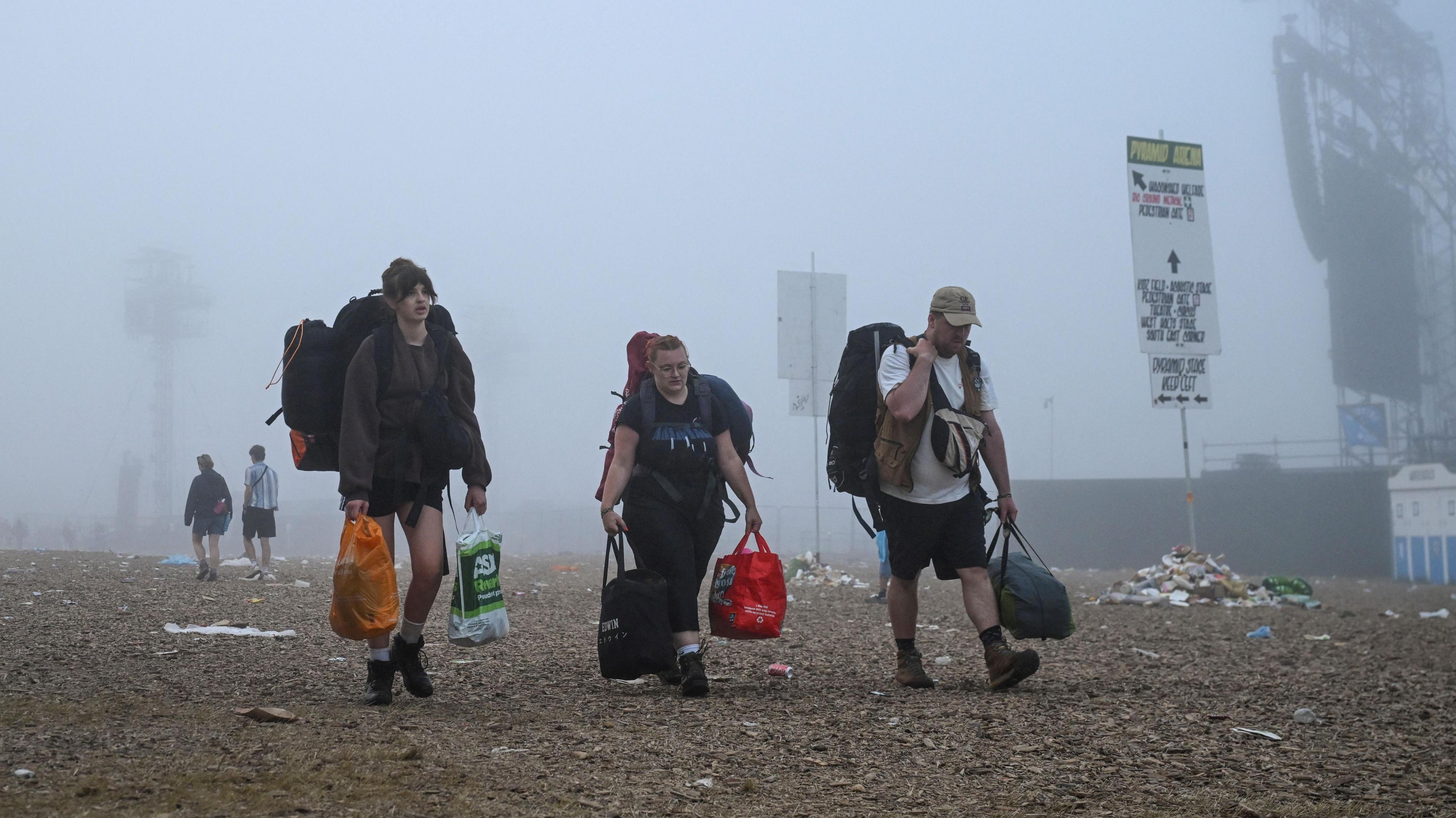 Three punters leaving Glastonbury Festival. They are all carrying bags and camping gear. There is mist around them and a few piles of rubbish scattered around signs.