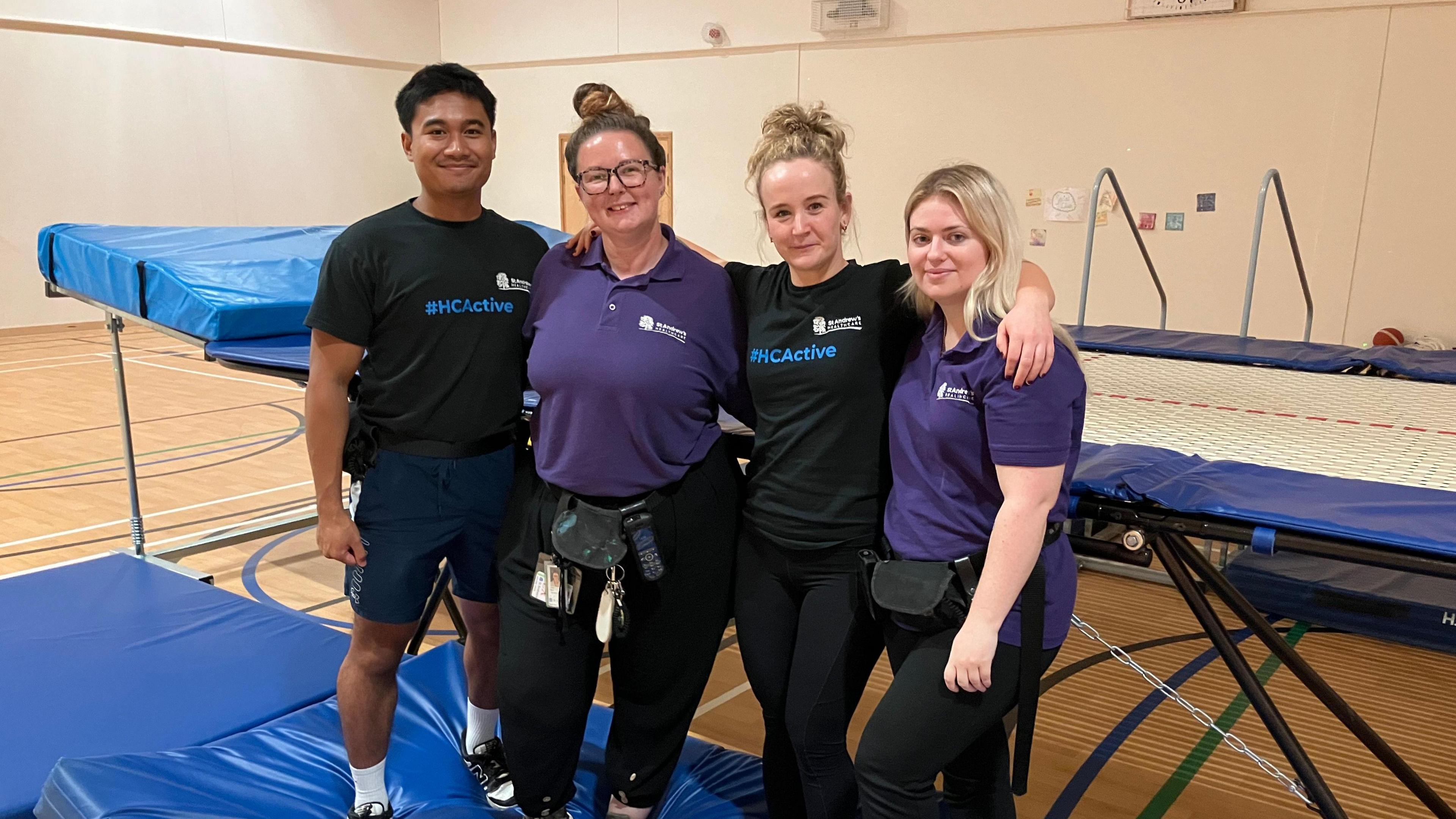 Four members of the rebound therapy team: a man in a black t-shirt and blue shorts, two women in purple polo shirts and black trousers, and a woman in a black t shirt and black leggings, stand next to a blue gymnastics trampoline in a sports hall. 