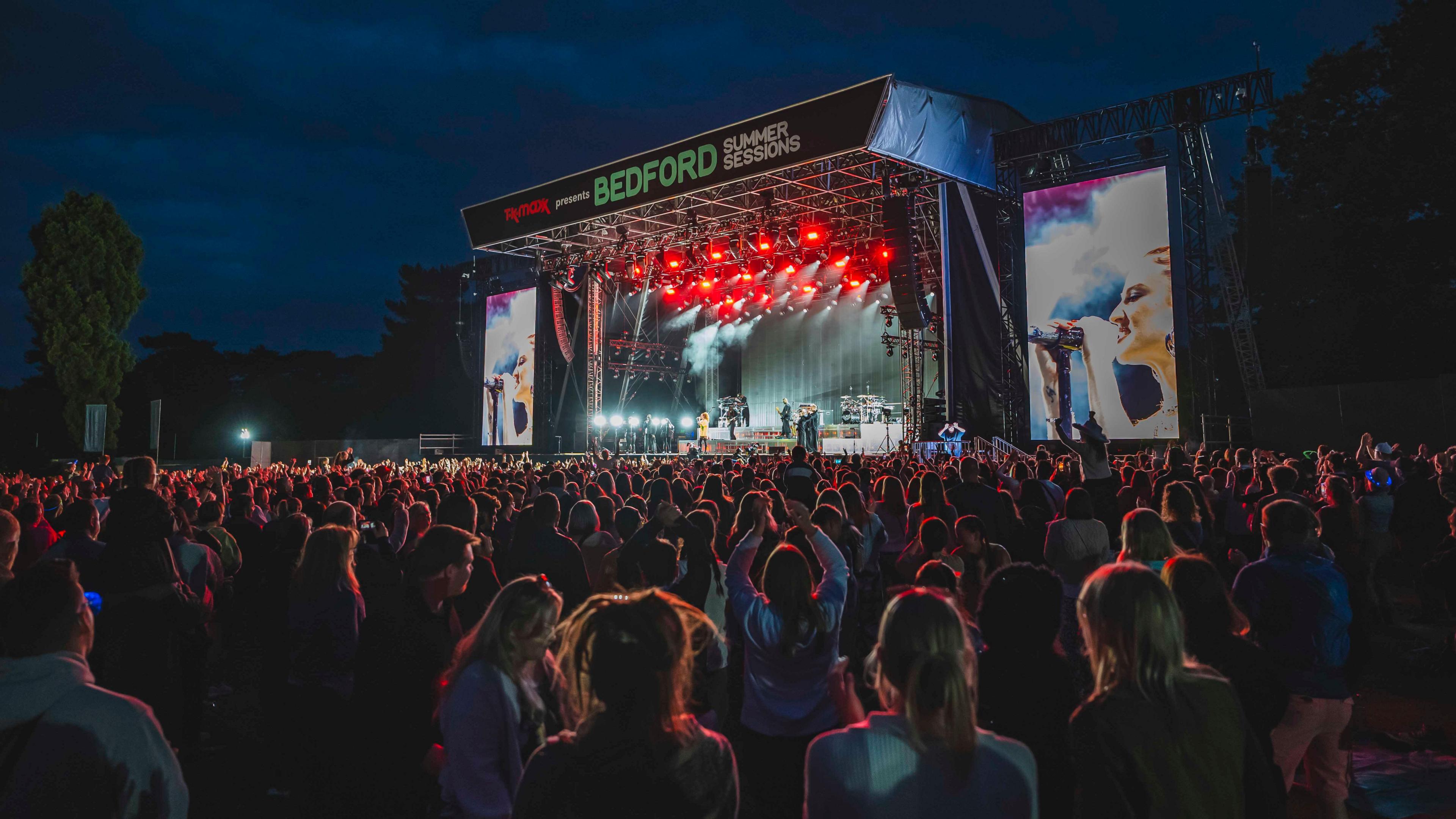 Jess Glynne performing in concert, showing audience members outside in a park, a large stage, with lights, projections, screens, a band and a dark sky. 