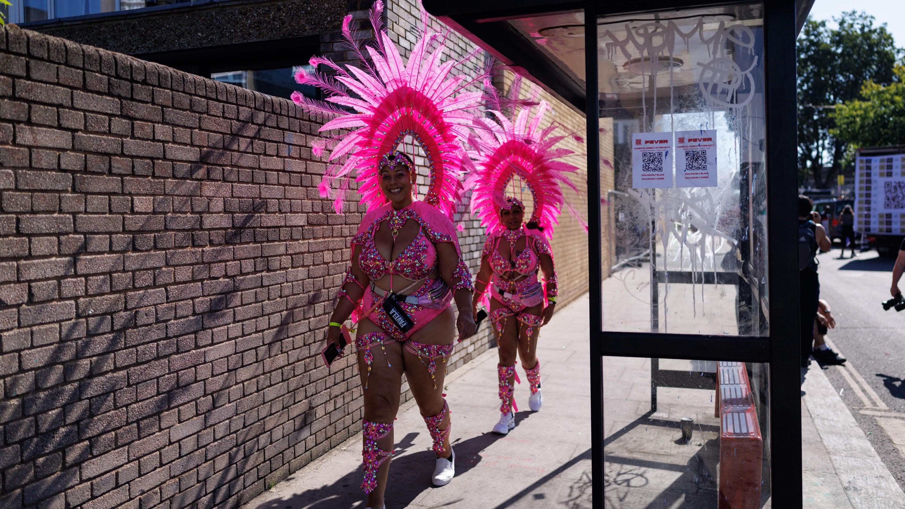 Two women in pink costumes walk past a bus stop on a street. Next to them is a brick wall.