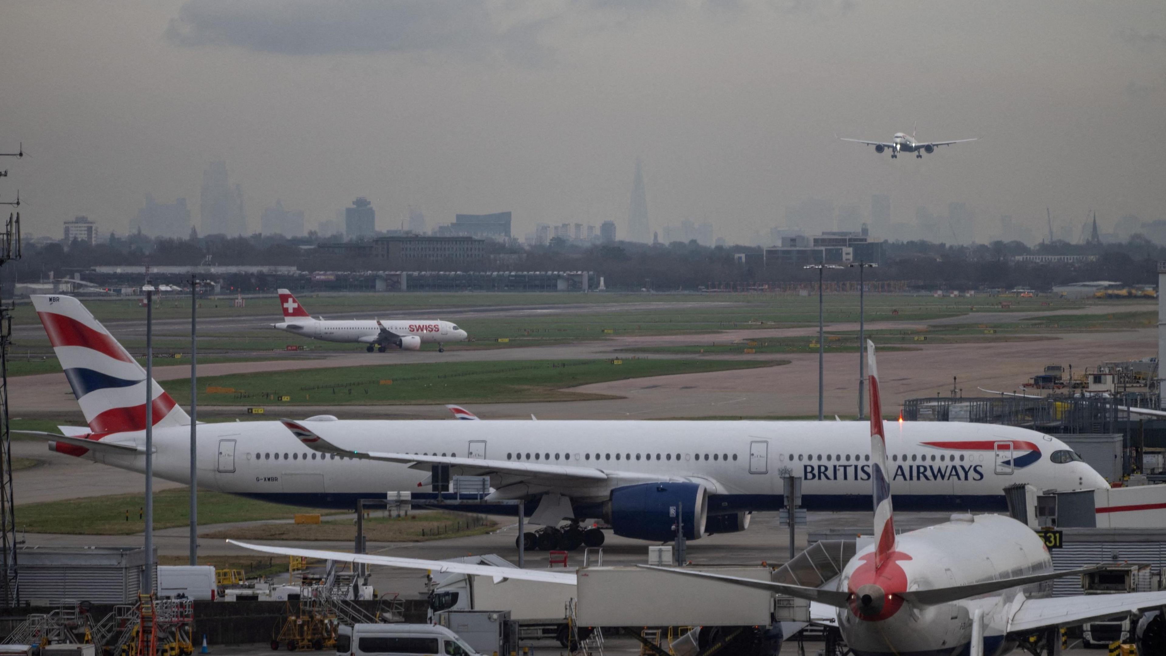 Planes arriving and departing from the runway at London Heathrow. One plane is coming into land, whilst others traverse the runways on the ground, where a BA plane parks at a gate whilst another smaller Swiss Airline plane also taxis behind it. The London skyline is clearly visible in the distance on a grey cloudy day, with the Shard being especially prominent.