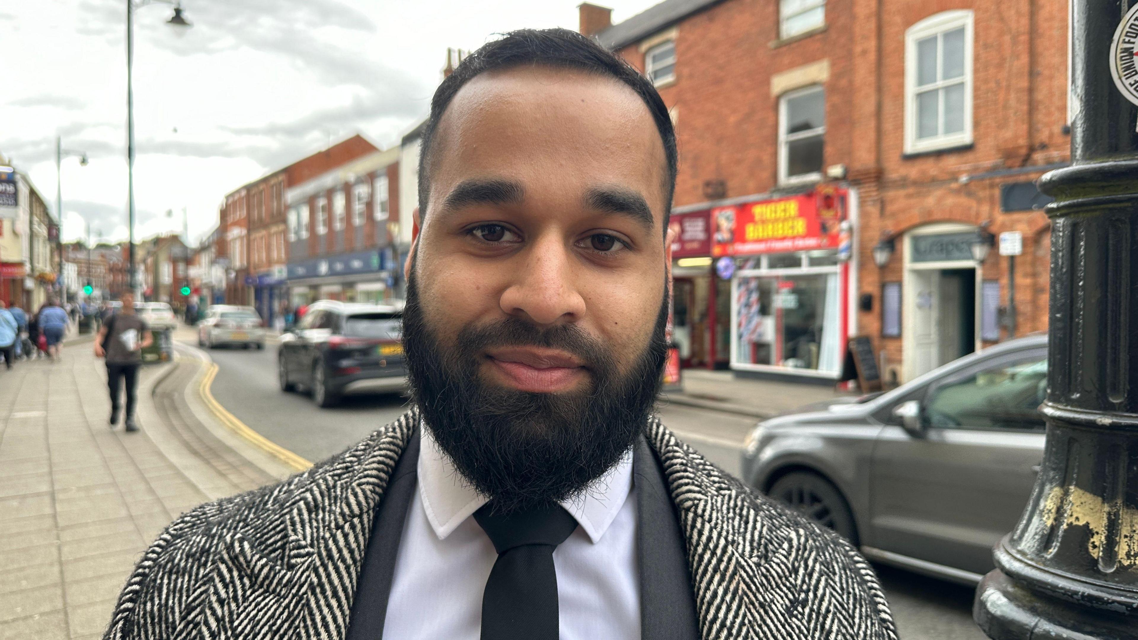 Ayaaz Ashraf posing for a picture in the street, wearing a black and white coat over a white shirt and a black suit jacket and tie. He is smartly presented with a beard