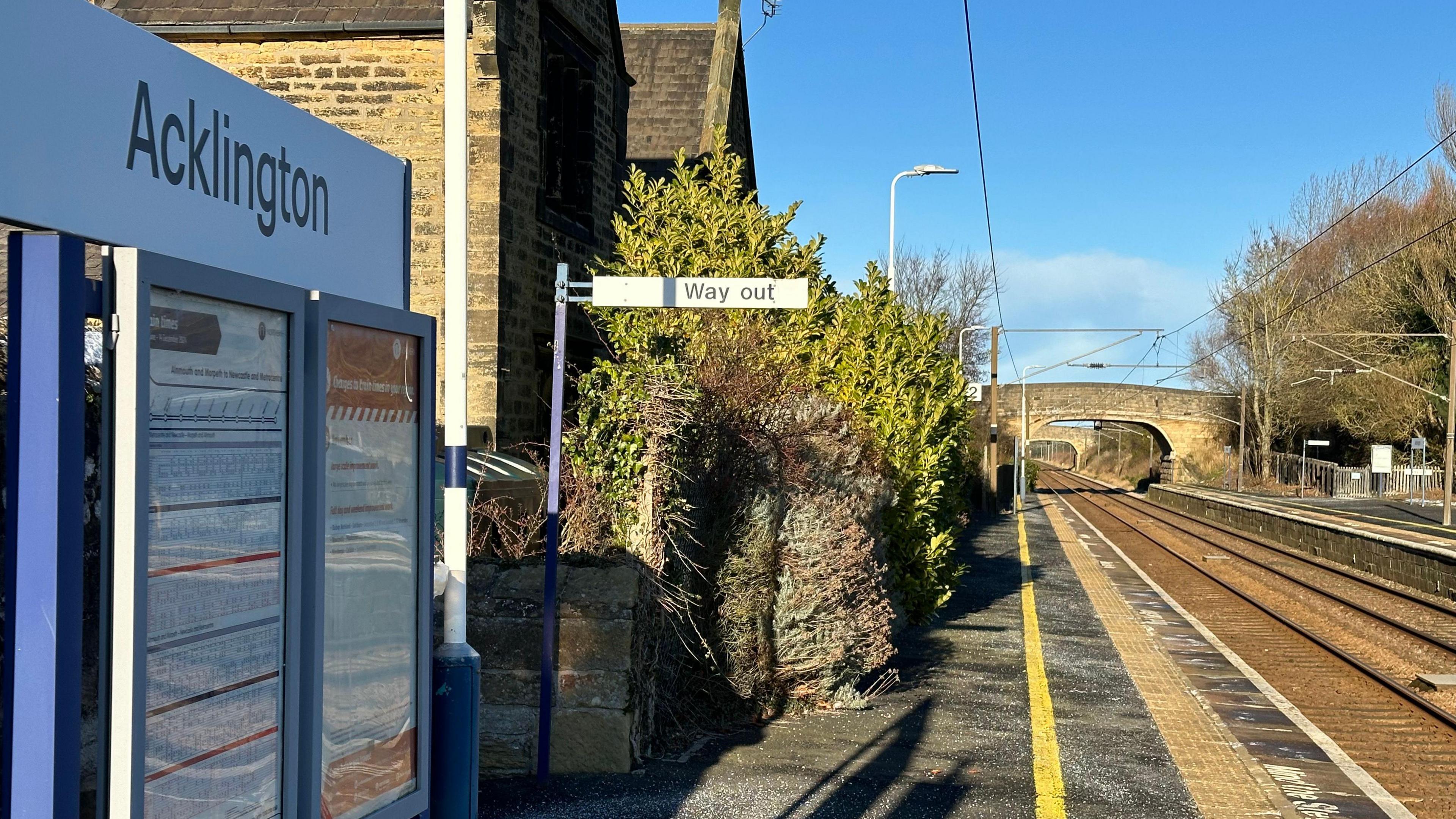 An empty platform in a very rural station with two bridges visible across the line ahead. A sign saying Acklington station is visible.