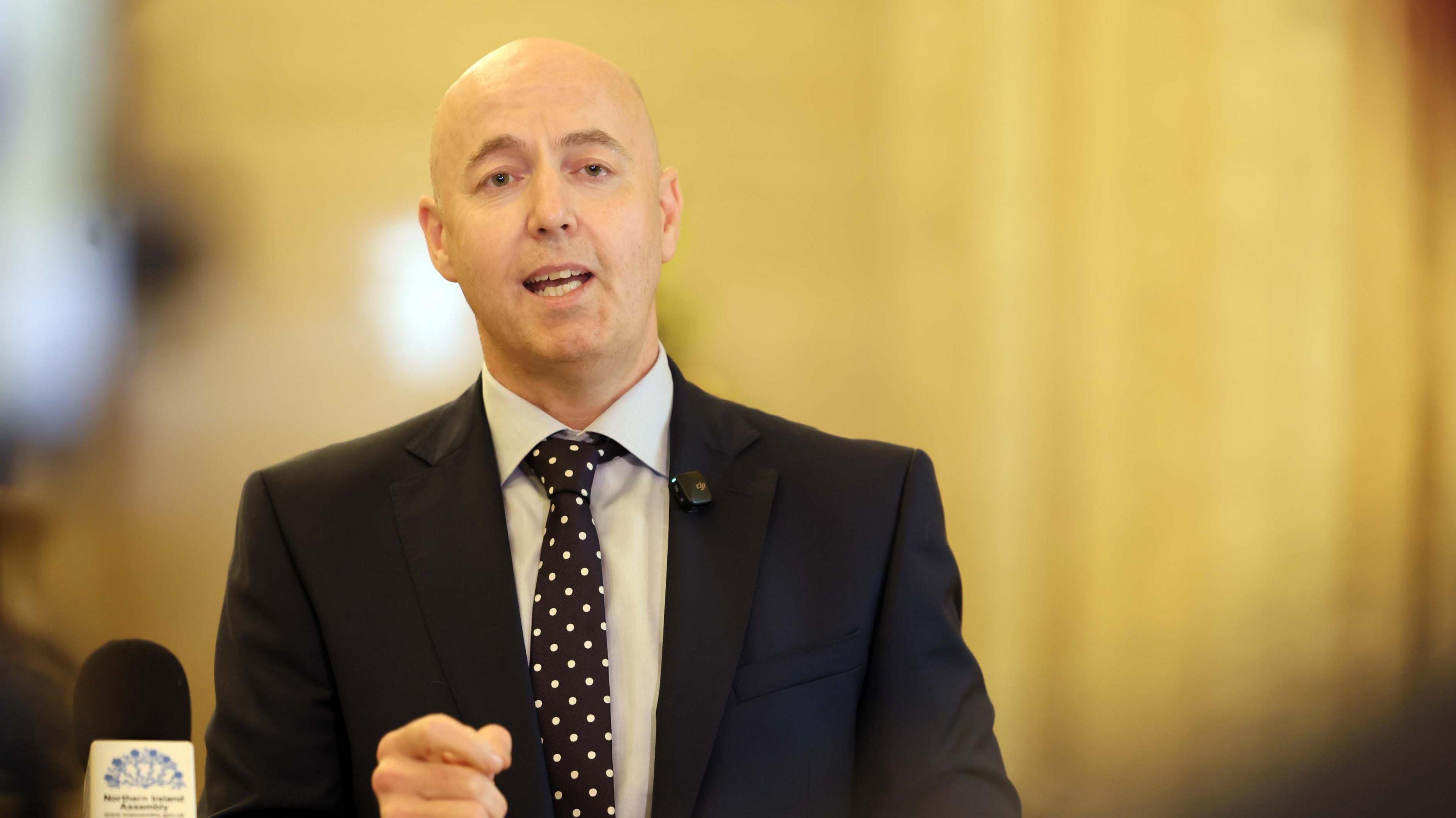 Burrows is wearing a suit and a spotty tie. A staircase is behind him. 
