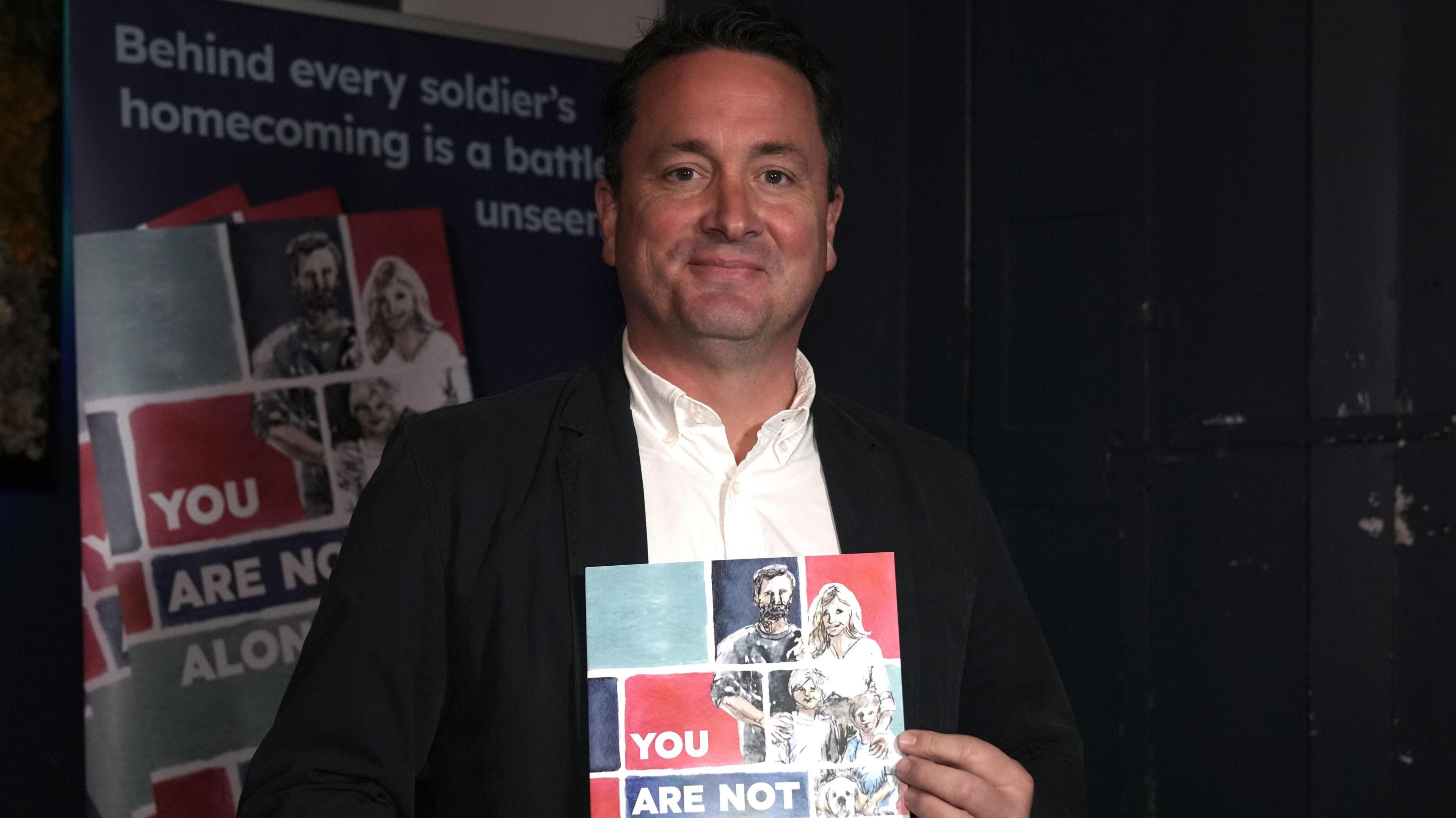 Rob Parker smiles at the camera as he holds his new book. He has short dark hair and wears a black suit jacket with a white shirt underneath. A poster promoting his book is behind him.