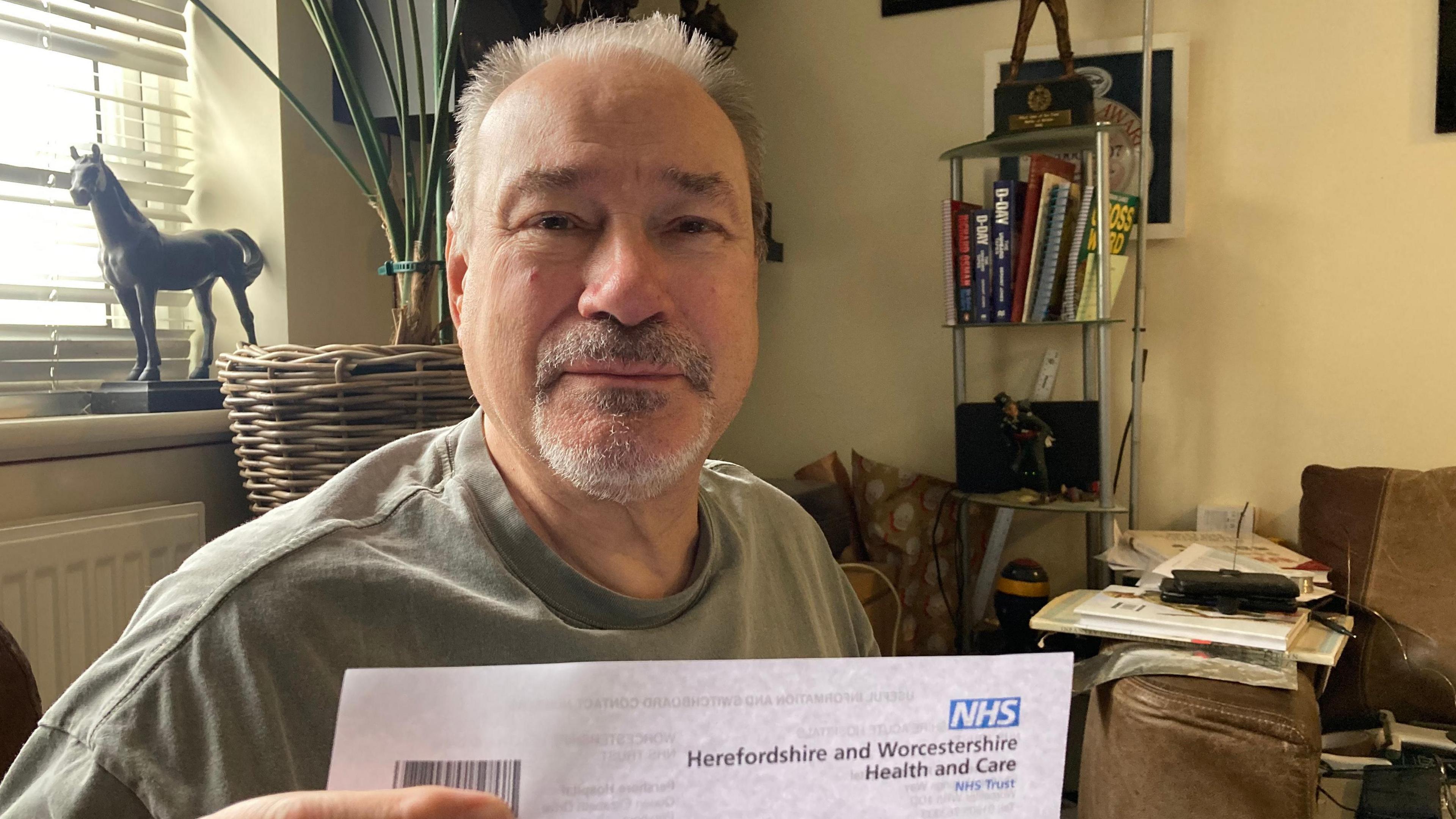 A man sat down on a sofa holding up a letter which has the NHS logo on it. He is wearing a green t-shirt and has a goatee. 