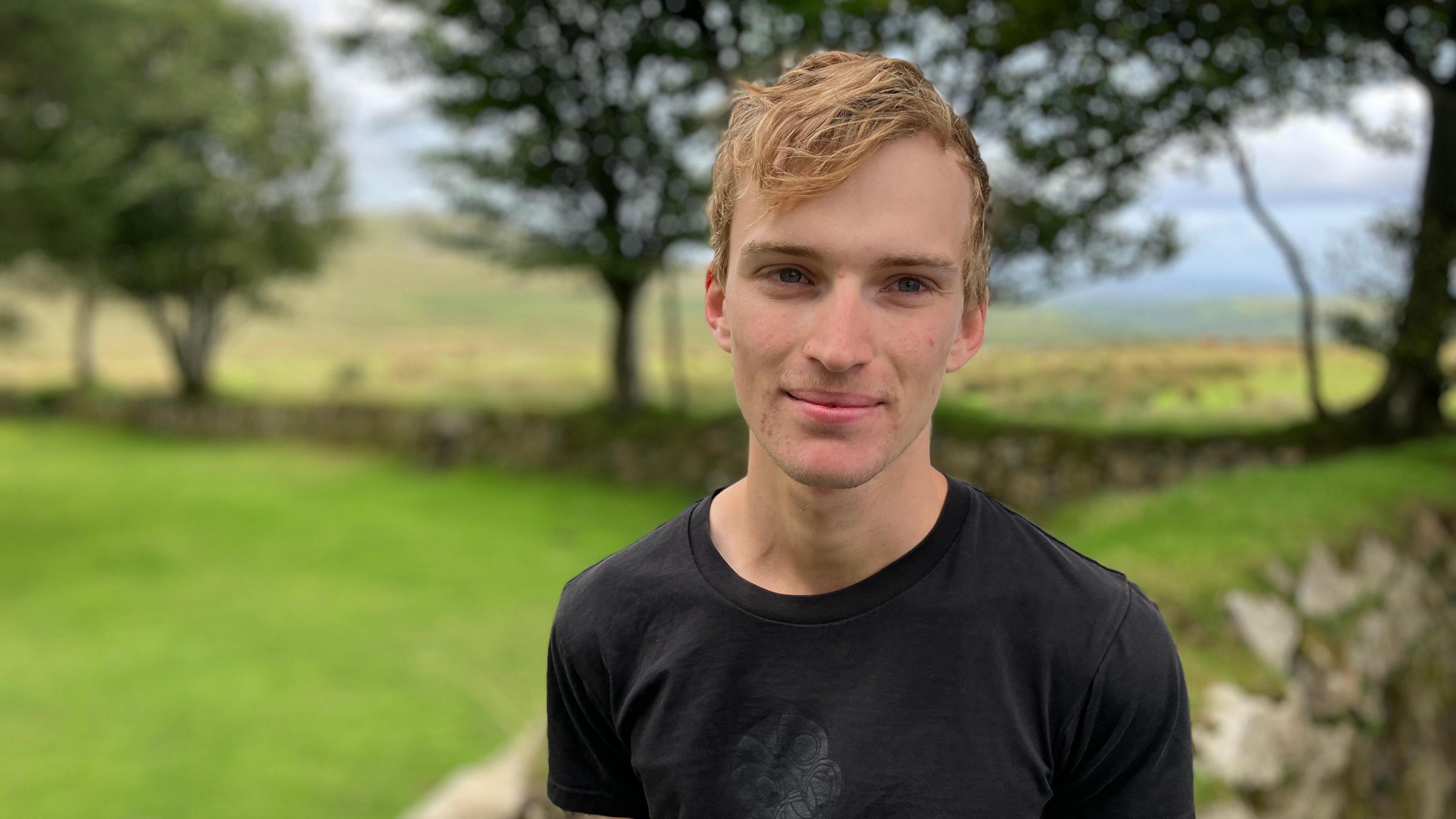 A man in a black t shirt stands in front of a stone wall on Dartmoor. There are trees and fields in the background.
