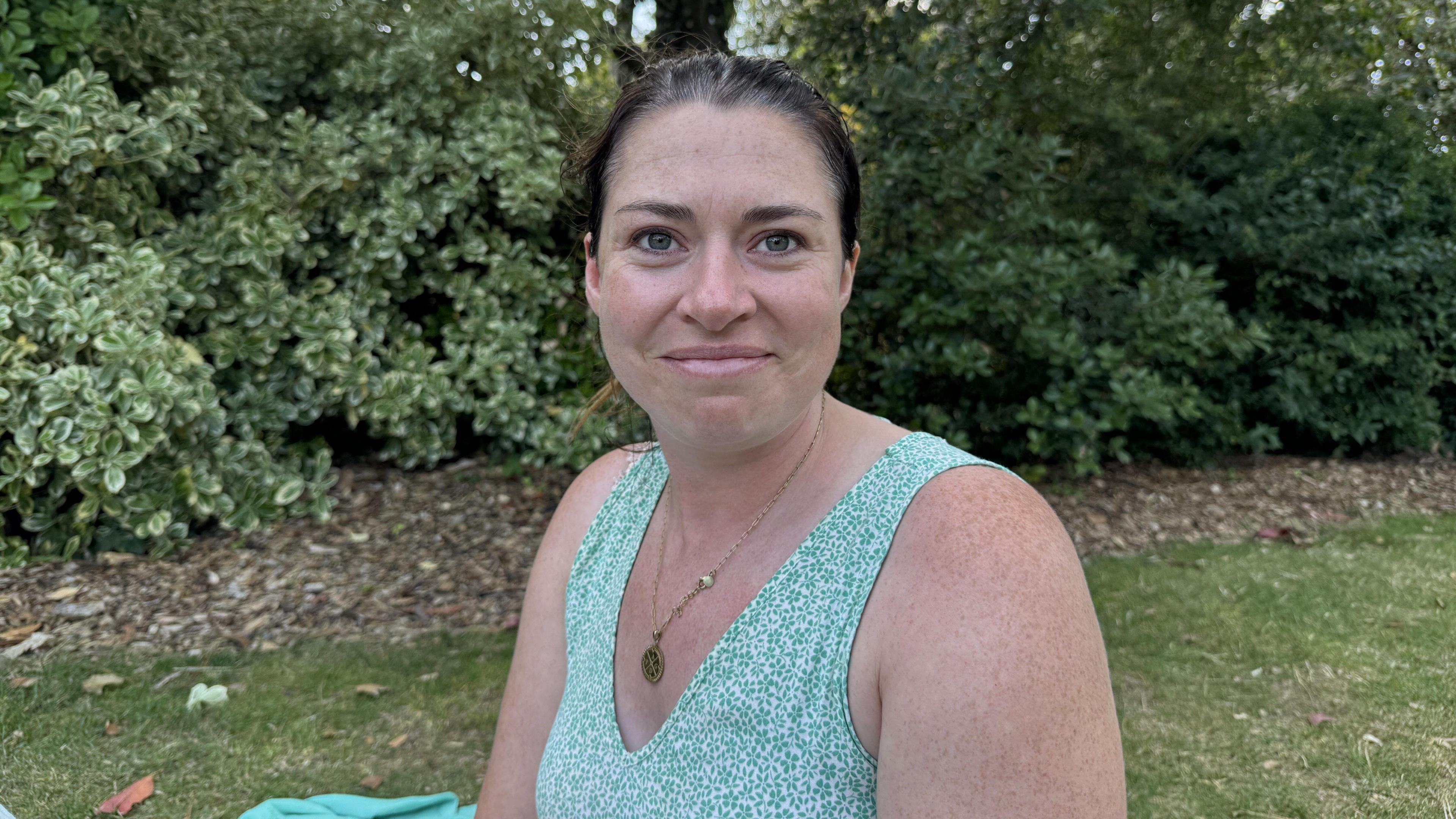 A woman sits on grass with green bushes behind her. She is wearing a sleeveless green patterned top and a delicate gold pendant necklace. Beside her lies a light green blanket. 