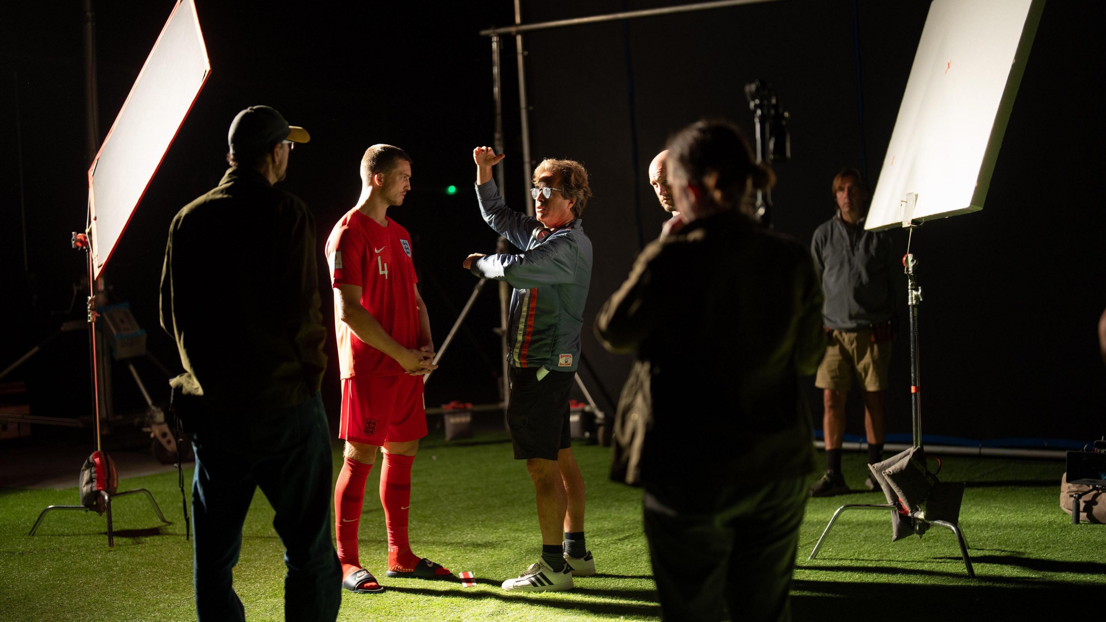 A director, wearing a blue jacket and shorts speaks to an actor wearing a red England men's football kit - they are standing on fake grass surrounded by production crew and lighting equipment