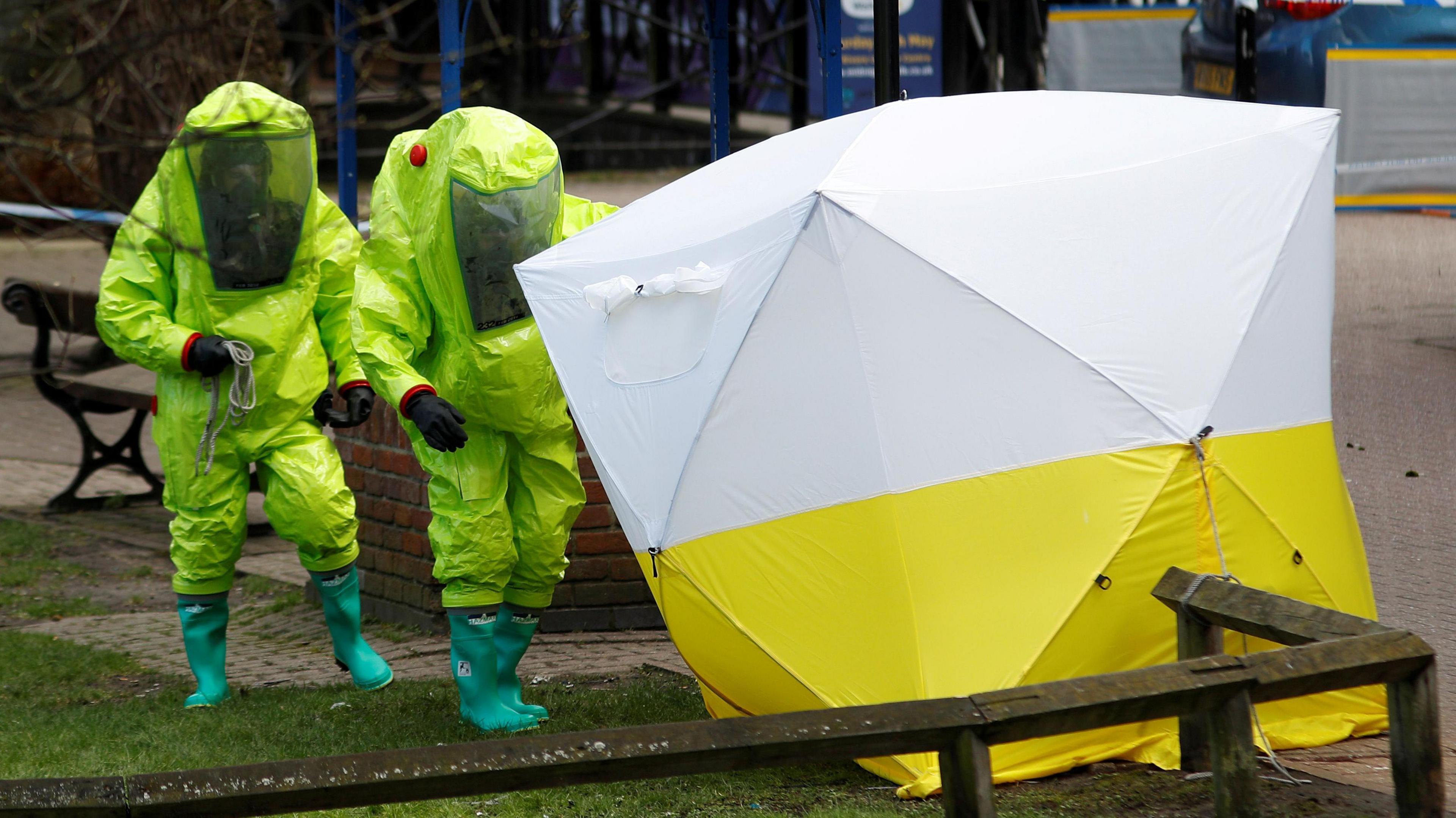 Two people in neon green/yellow hazmat suits approach a large yellow and white forensics tent at the site of the poisoning. 