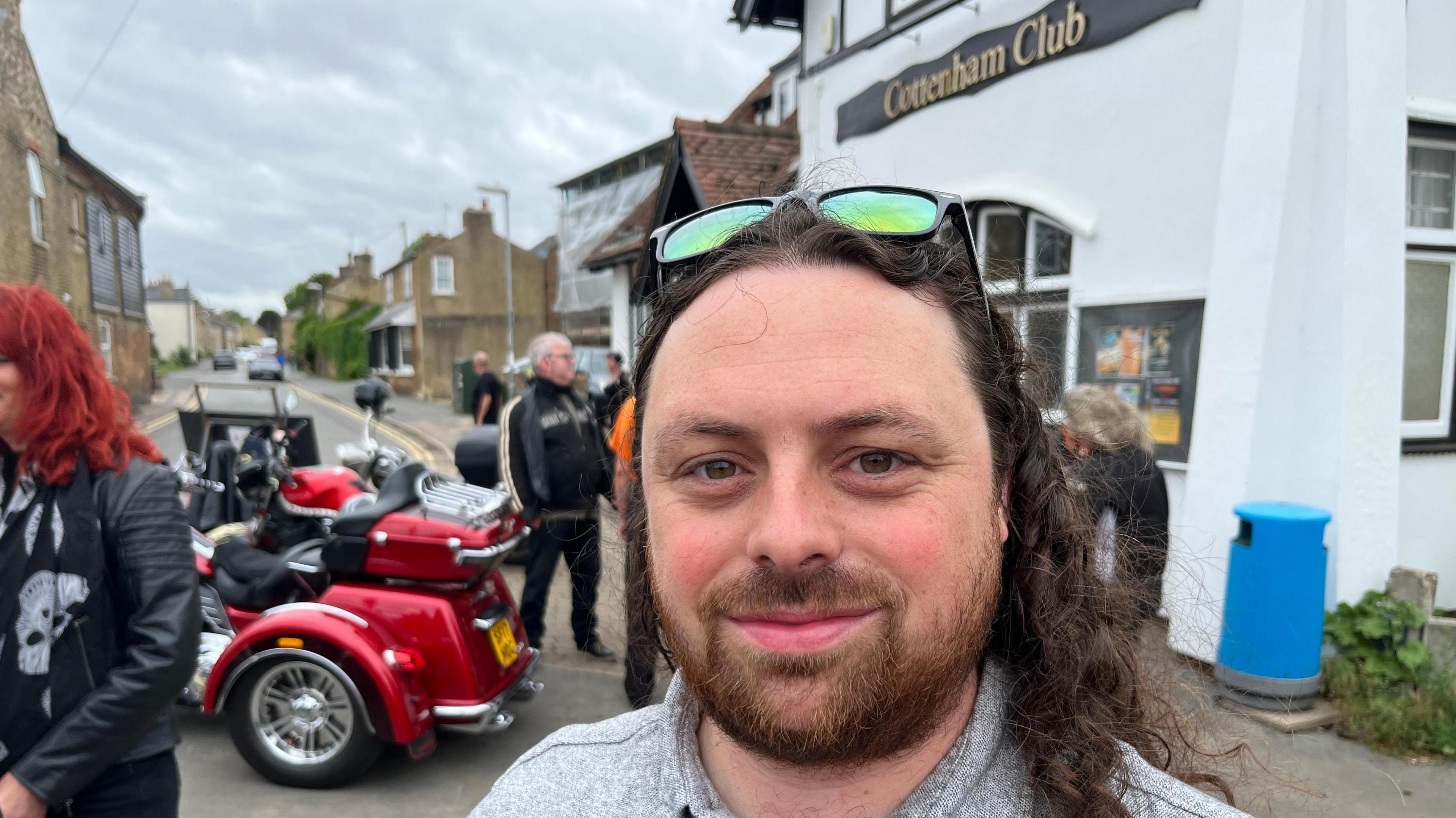 Dan Phillips is looking at the camera. He has long, wavy brown hair and a beard and moustache. He is wearing a grey top. There are people and a large red bike behind him