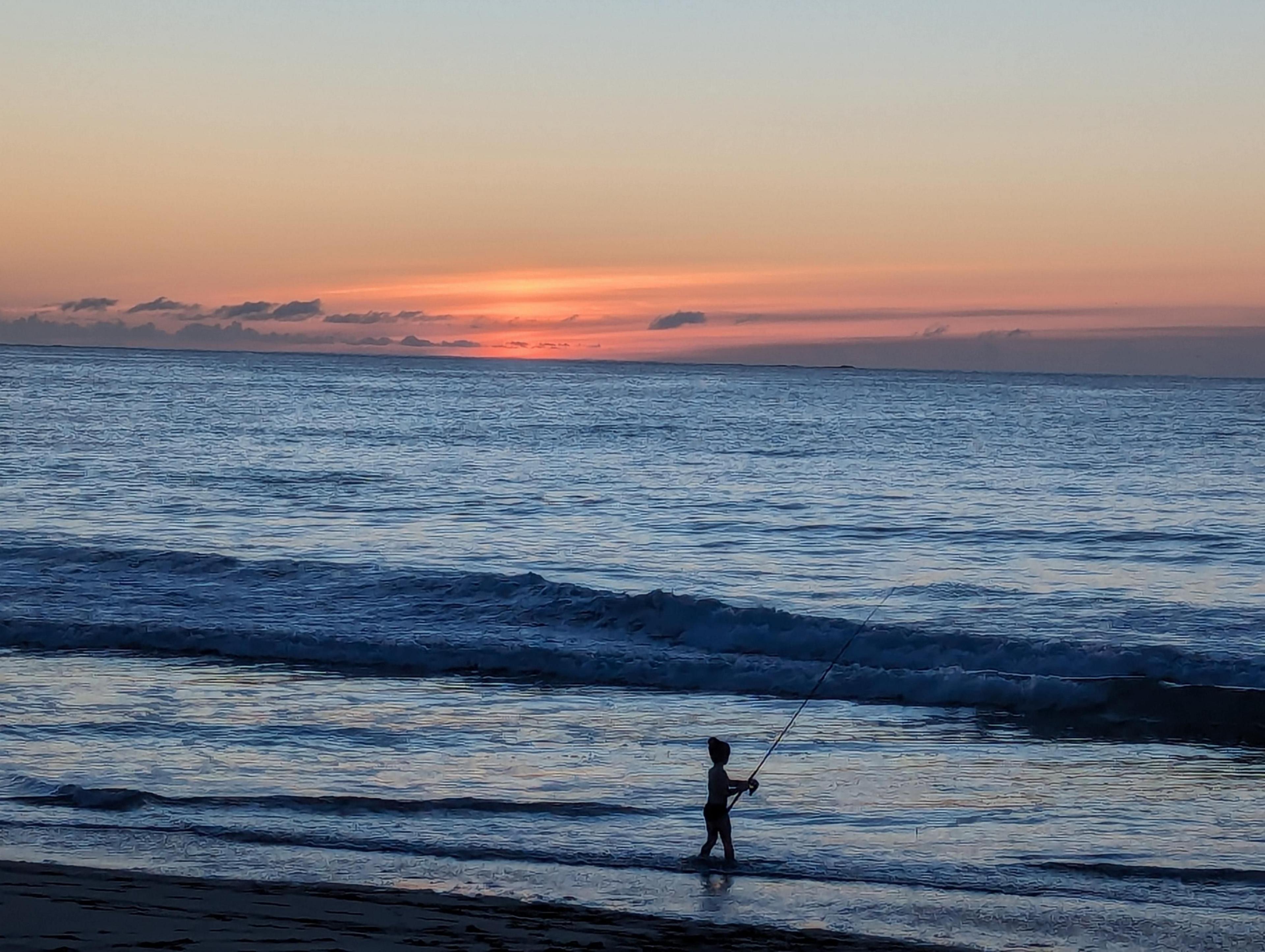 Young boy standing in shallow water at sunset on the beach, fishing.