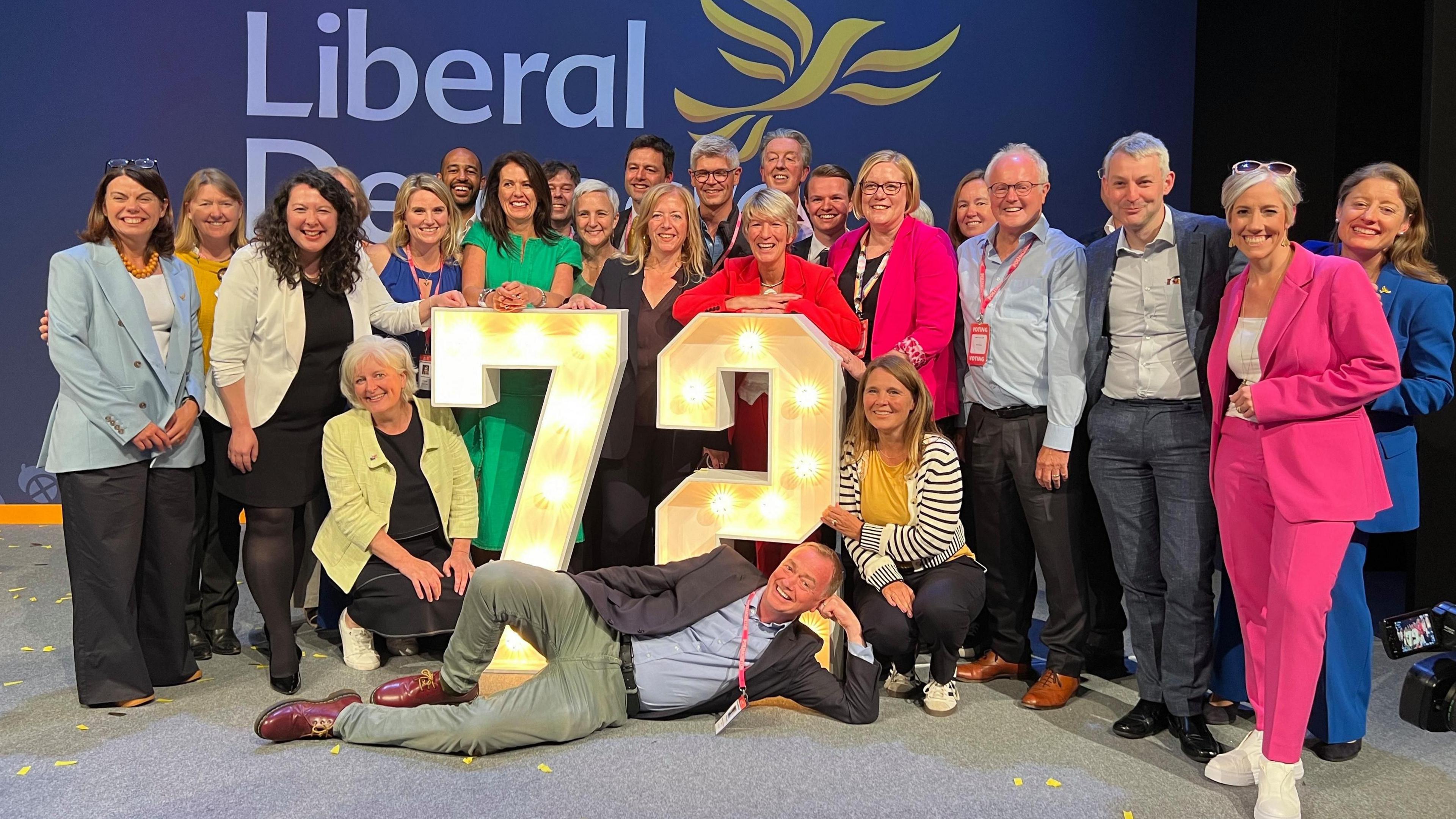 Liberal Democrat MPs pose happily around a giant 72 sign, made of lightbulbs 