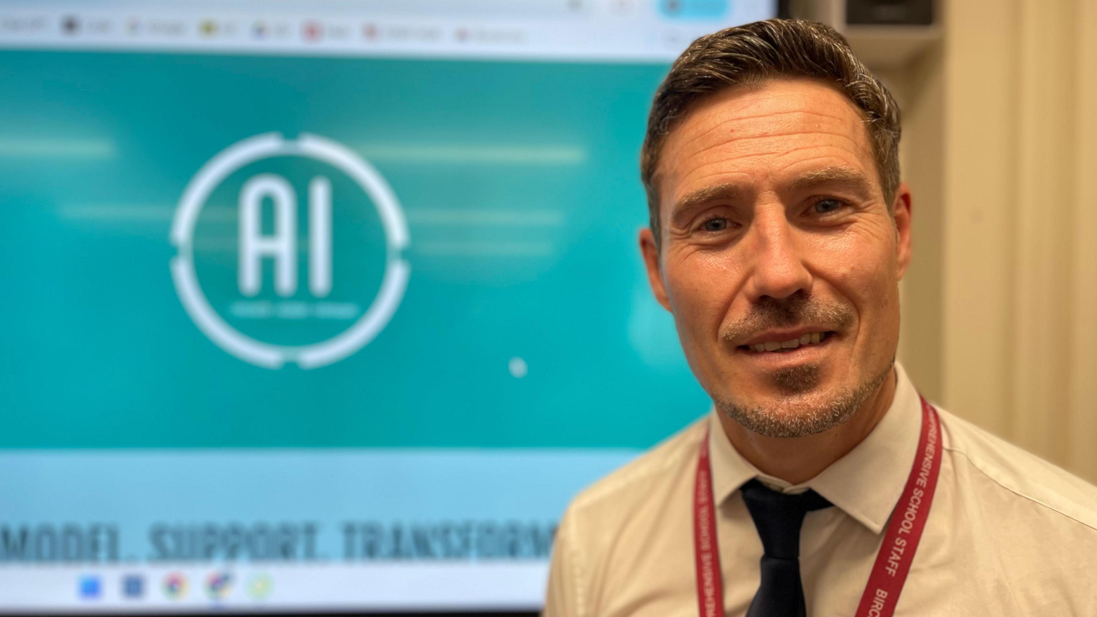 A teacher in a white shirt with a red lanyard is standing in front of a blue screen with the letters AI circled in white on it.