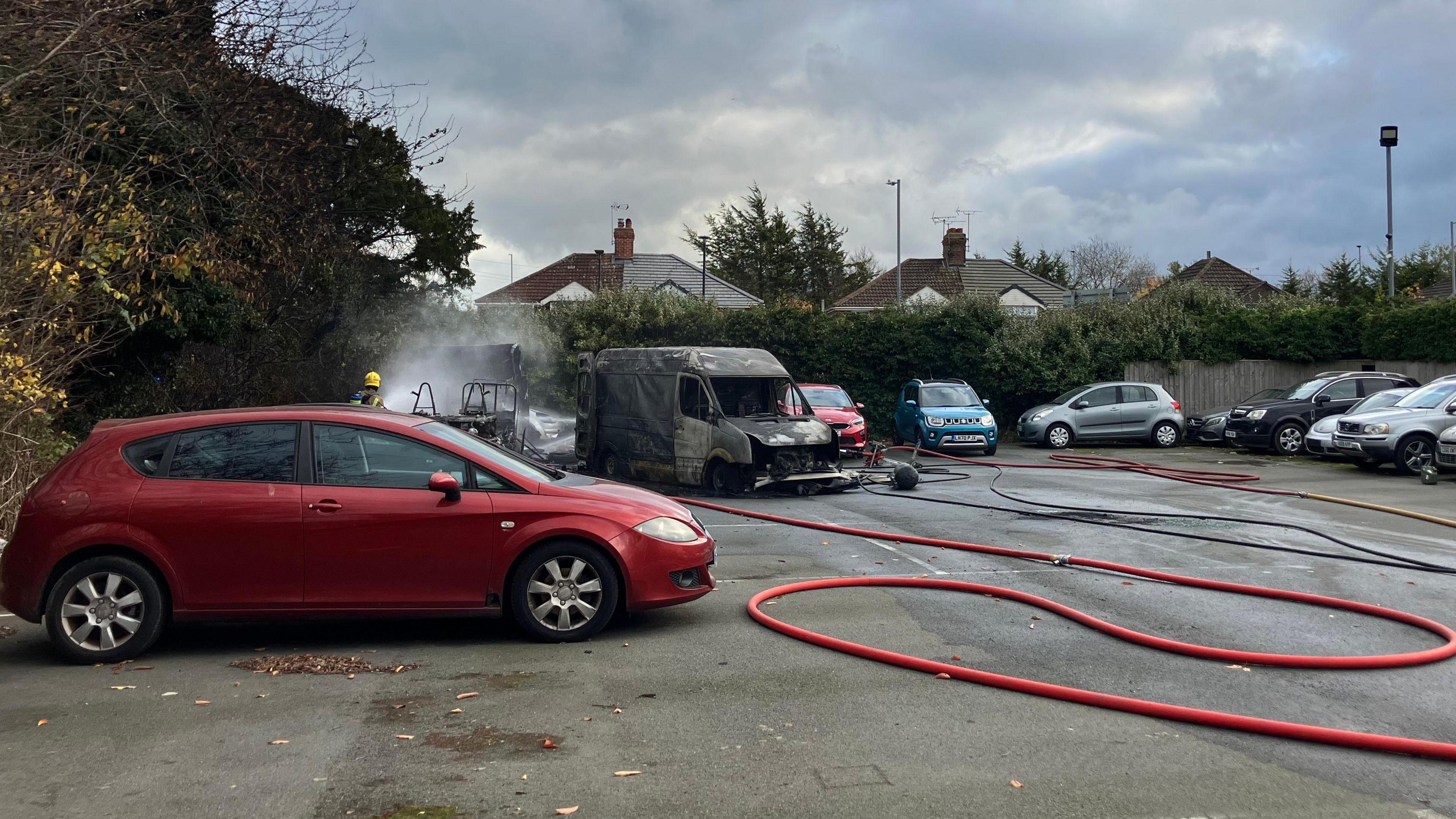 A burned out van in a car park with a long red firehose snaked out in front of it. Various other cars including a red Seat and a grey Volvo can be seen parked in the background.