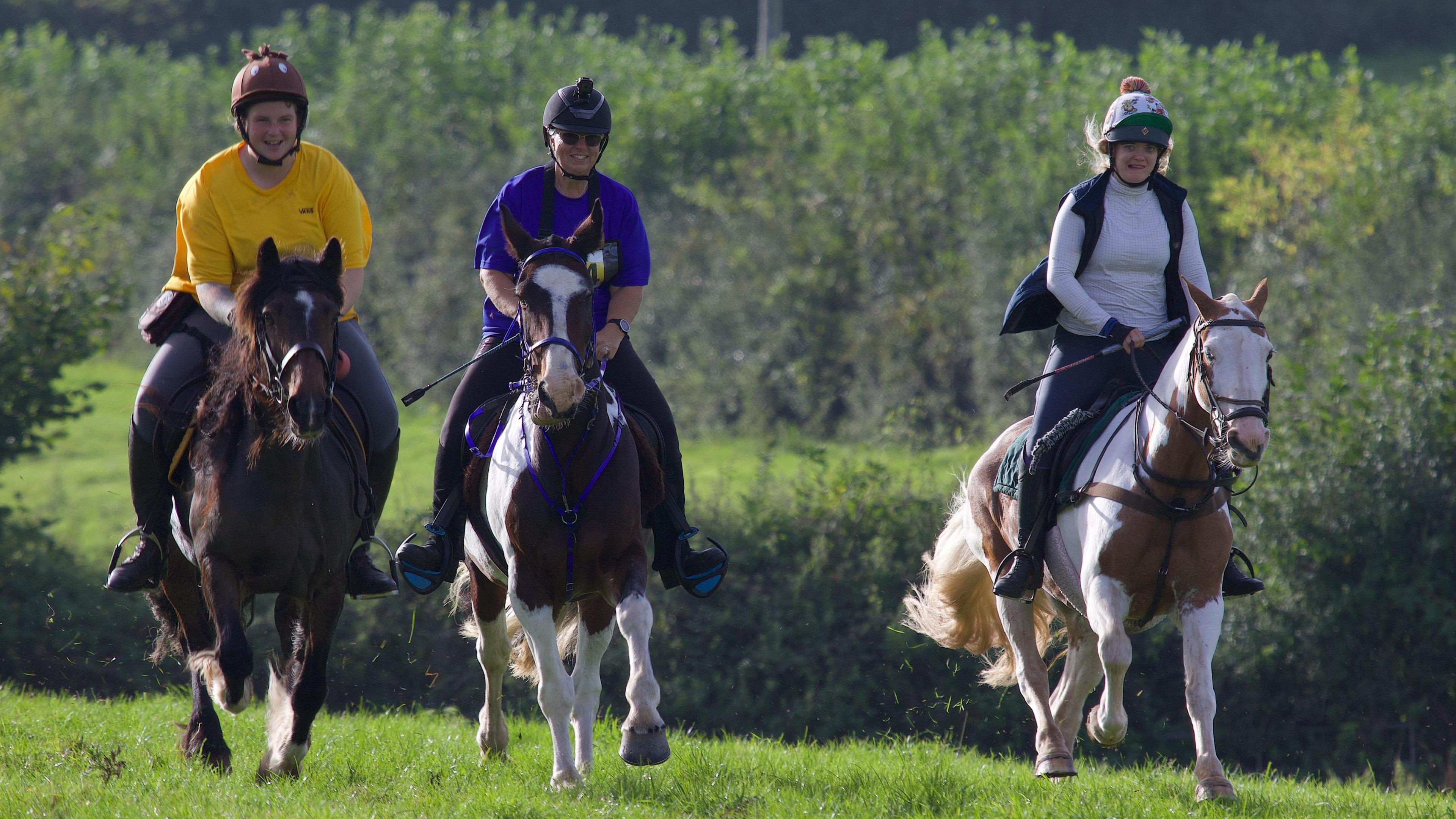 Three people sit on horses, all of them moving quickly towards the photographer with smiles on their faces. All are wearing helmets, and green fields can be seen behind them.