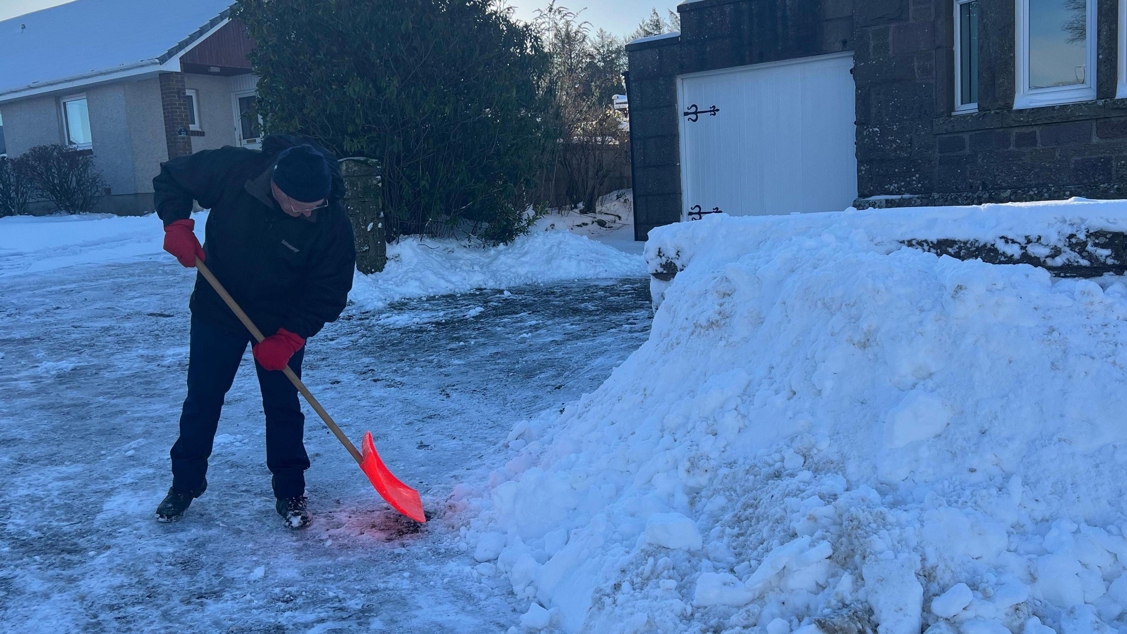Një burrë pastron borën me lopatë në anë të një rruge të akullt. Ai mban veshur doreza dhe rroba të ngrohta të papërshkueshme nga uji.