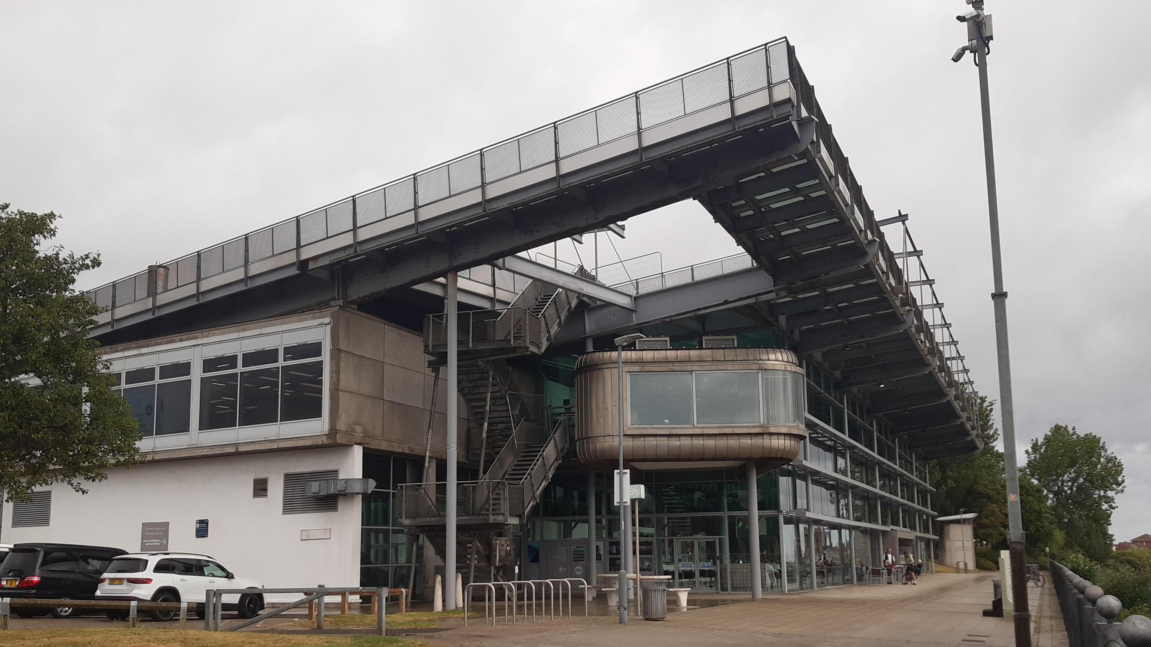 The National Glass Centre is a modern building made mostly of grey panels and glass. It sits next to the River Wear and there is a cafe seating area outside. 