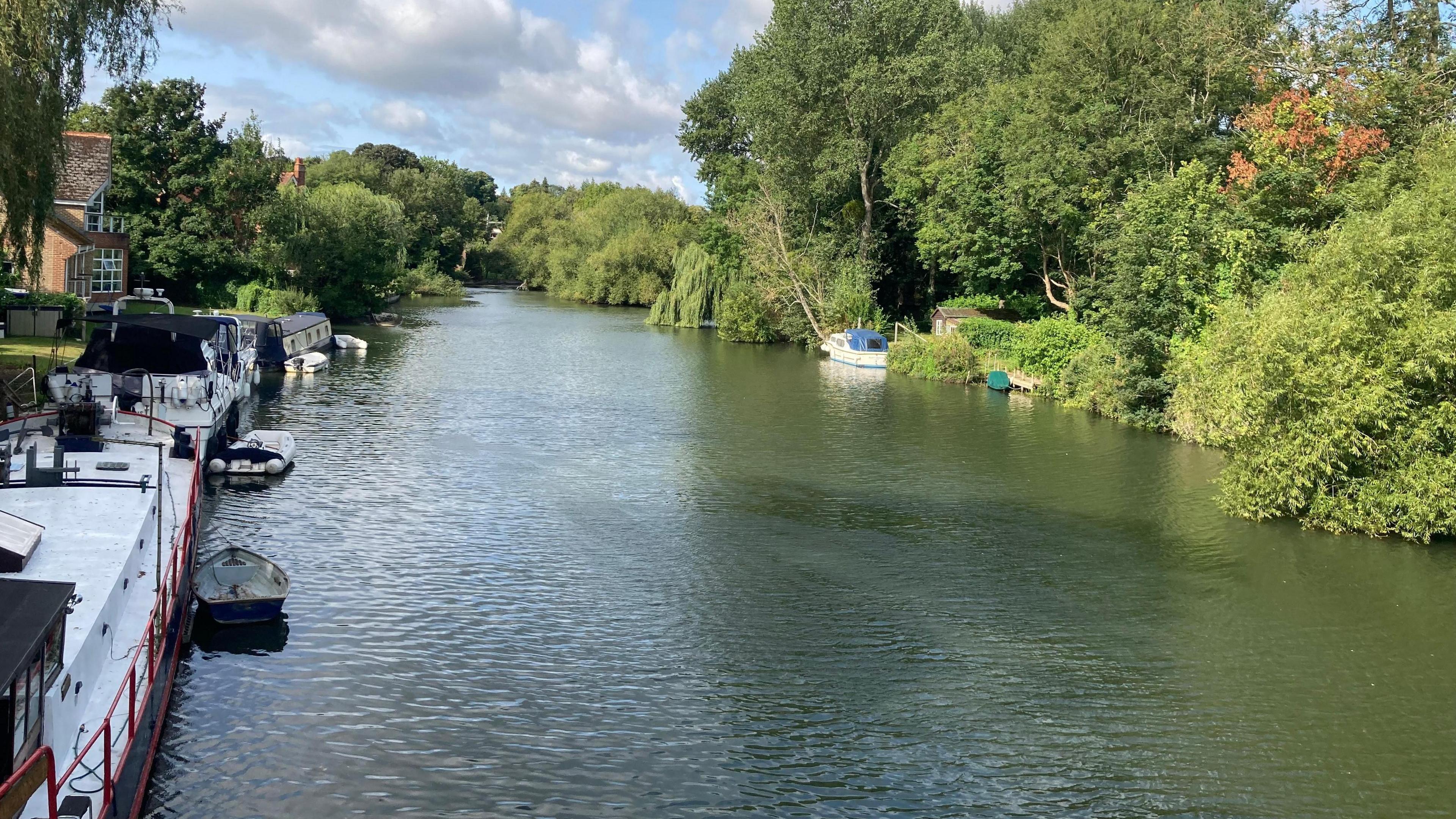 The river at Pangbourne on a sunny day, there are white boats moored on each bank. On one side of the river are green trees, and on the other side you can see some brick homes.