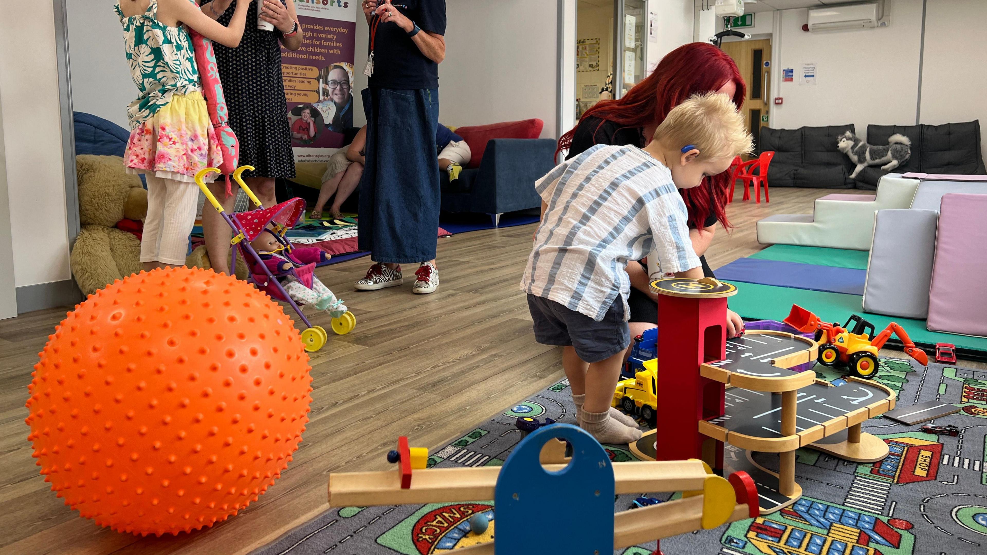 A small child with blonde hair, wearing a blue and white striped top, blue shorts and a blue hearing aid/cochlear implant can be seen playing with toy cars on a large play mat, with a woman with long red hair. Other people can be seen in the background, including a child and two adults, but their heads are not visible in the picture. A large orange ball is also visible in the front of the photo. 