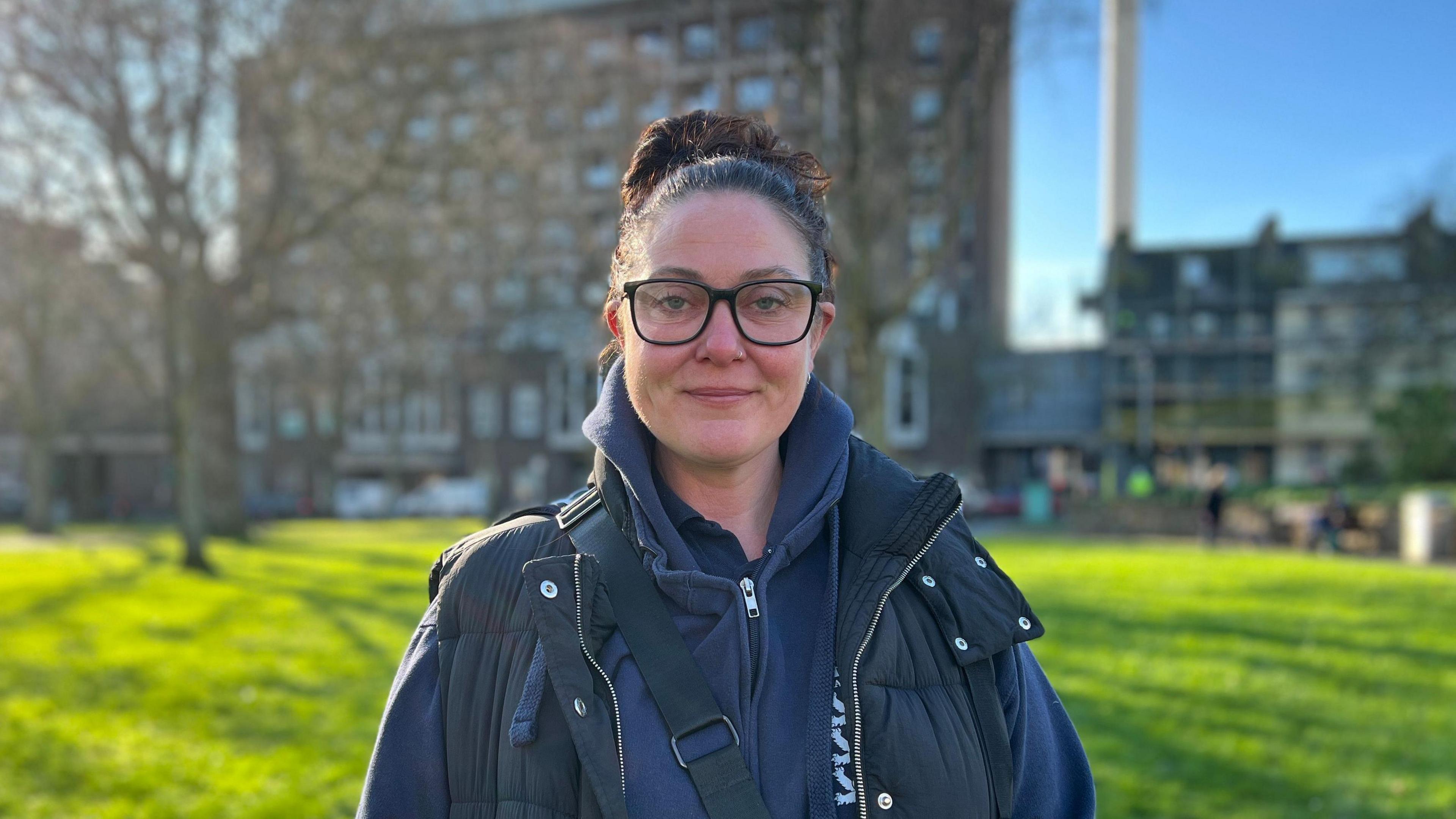 A picture of Sarah De Gruchy in a green space in Jersey. She is wearing black glasses and has her hair tied up.