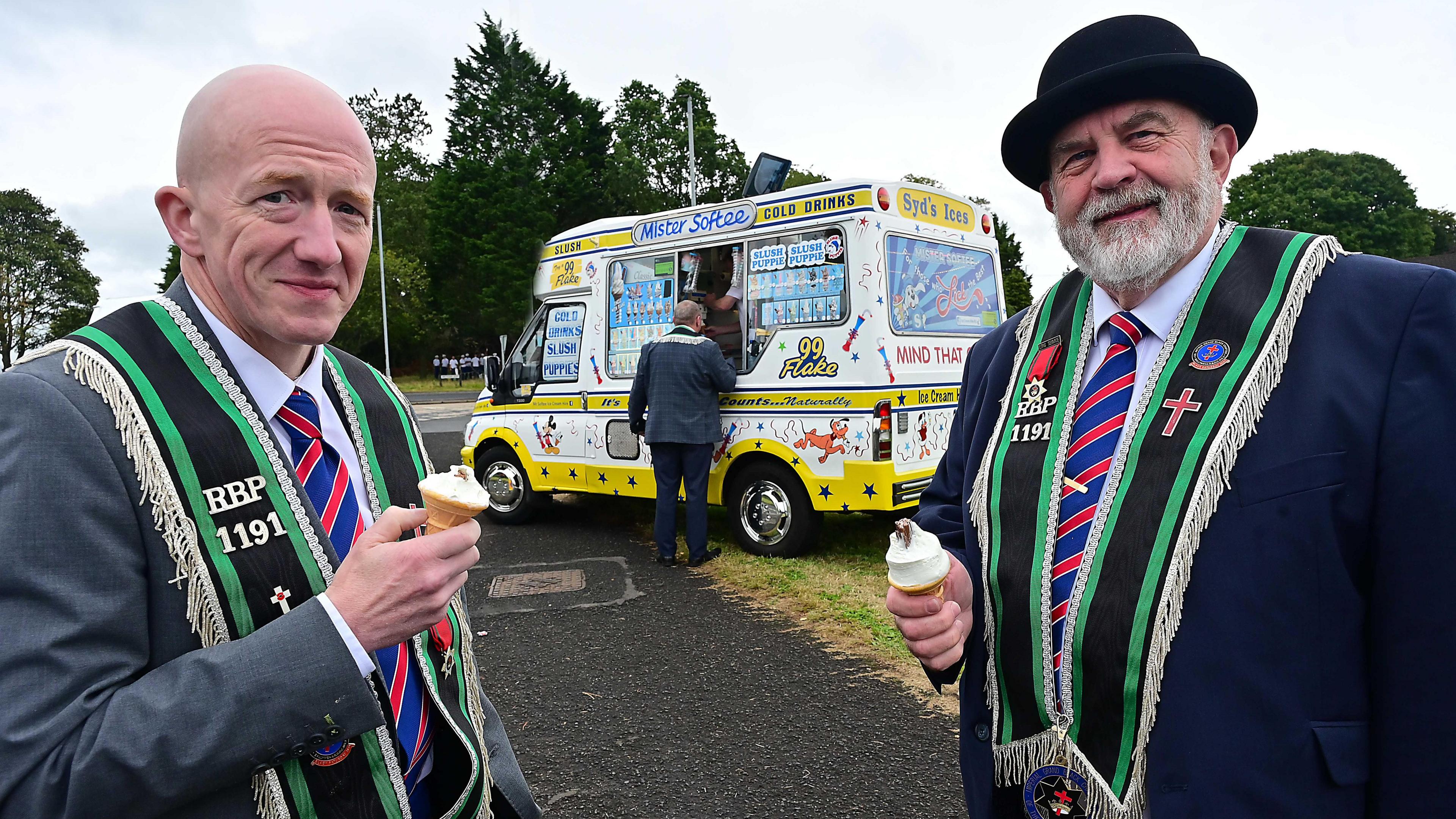 Two men with Mr Whippy ice creams standing in front of an ice cream van. They are wearing dark suits and black sashes with green trim, the man on the right has a black bowler on and the man on the left is bald.