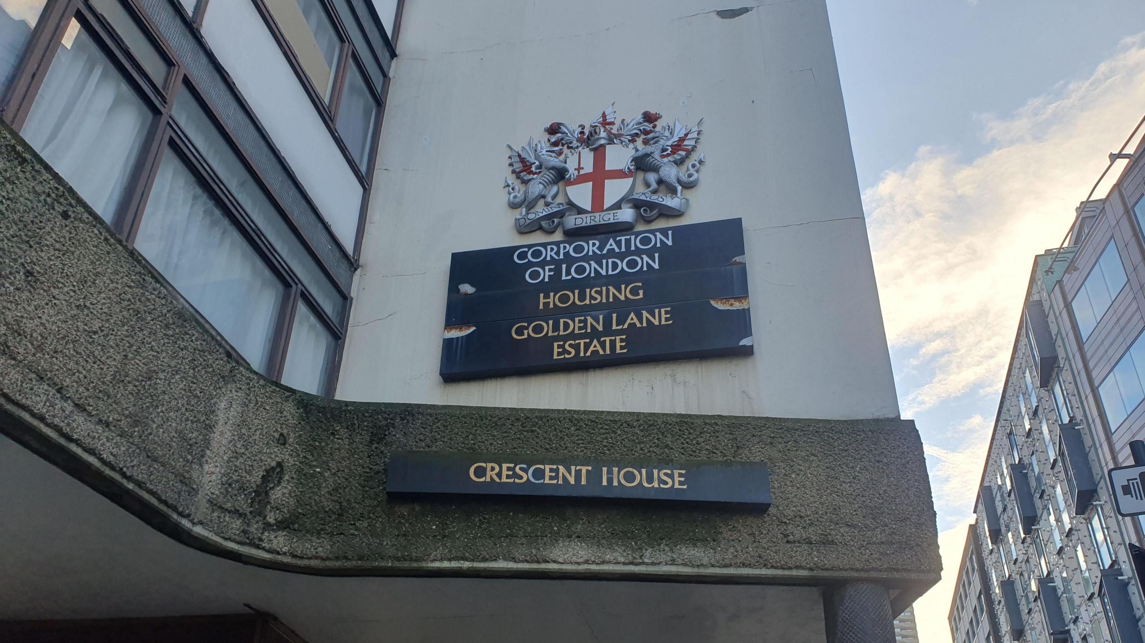 A close up of signage of Crescent House. Signs display the City of London's crest and the words “Corporation of London," "Housing," "Golden Lane Estate,” and below that "Crescent House".