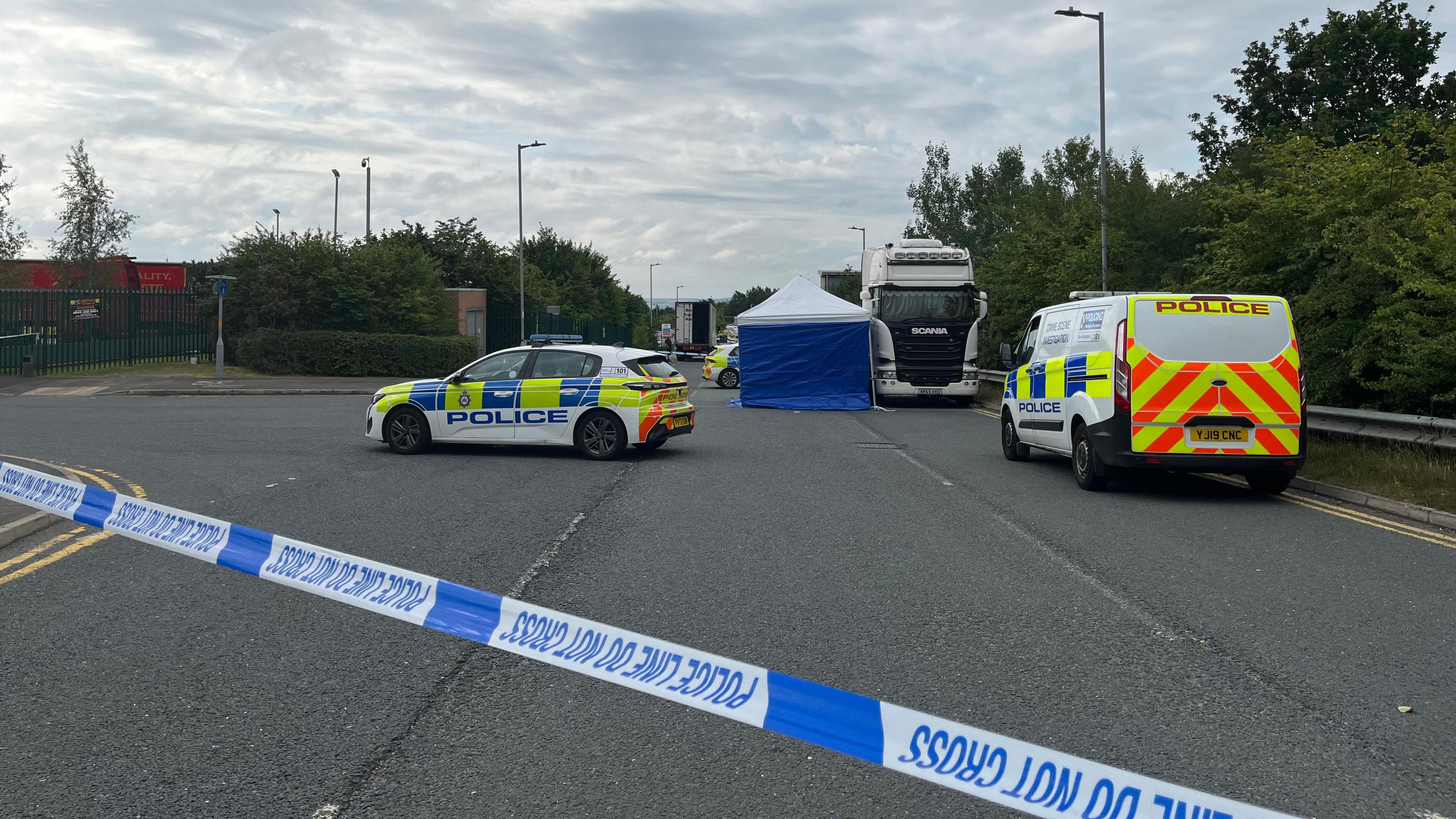 A police forensic tent stands next to a lorry at the side of the road. There are two police cars and a van alongside, with police tape in the foreground.