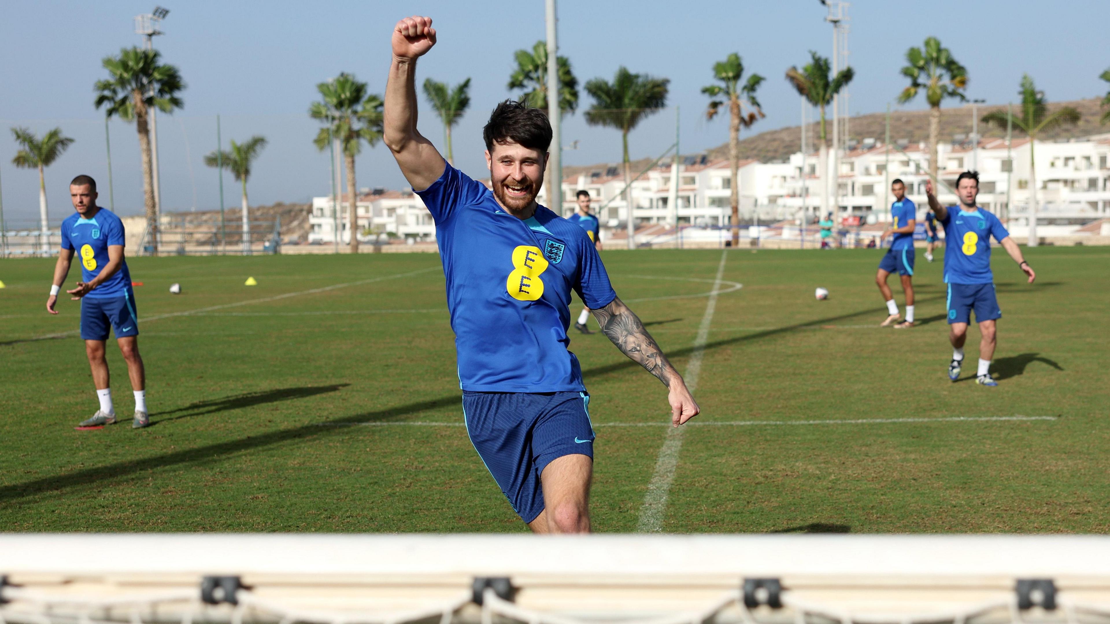 Harry Baker training in a blue England shirt at a training camp in Spain in January 2024. Teammates are training alongside him and he is raising his right fist in the air, as if celebrating, and smiling