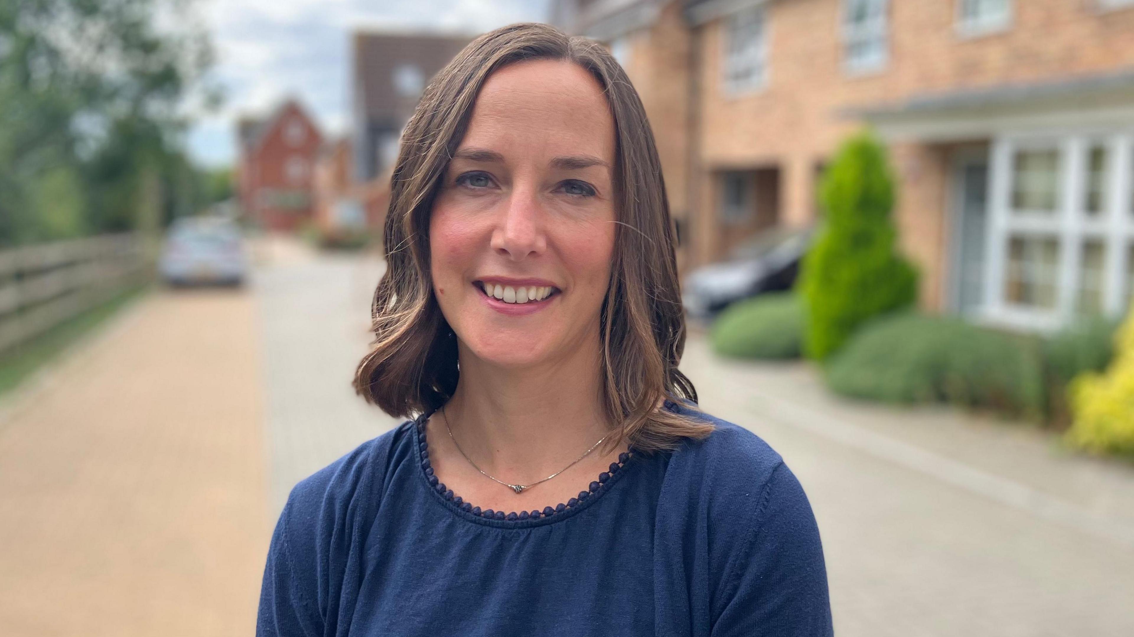 A head and shoulders picture of Dr Keetie Roelen who is smiling at the camera and wearing a navy blue top. The background to the picture is blurred but she is standing on a residential street with homes on one side of a road and a fence and some green trees and bushes on the other side.
