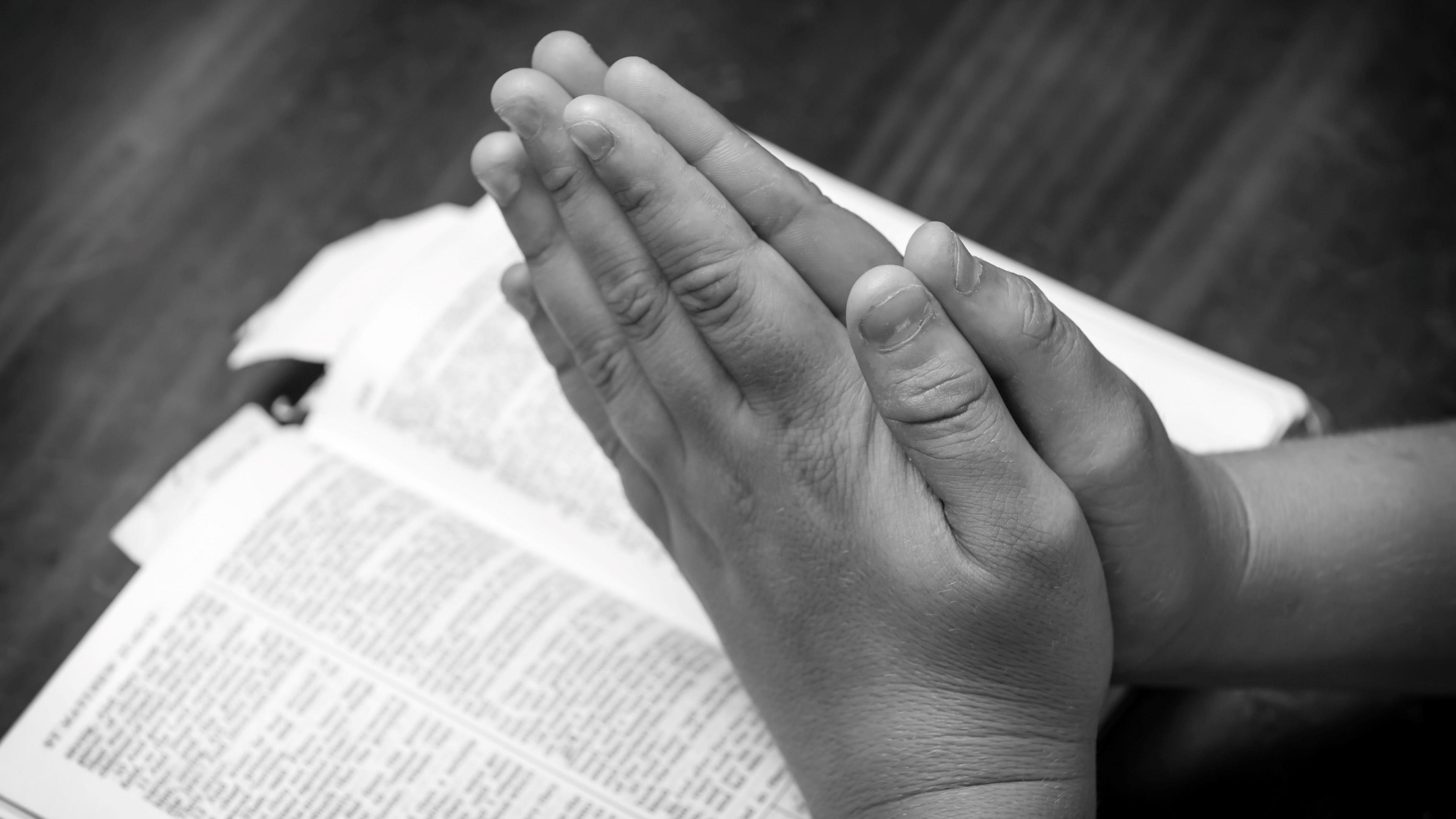 A child's hands are joined in prayer. A bible lies on a desk.