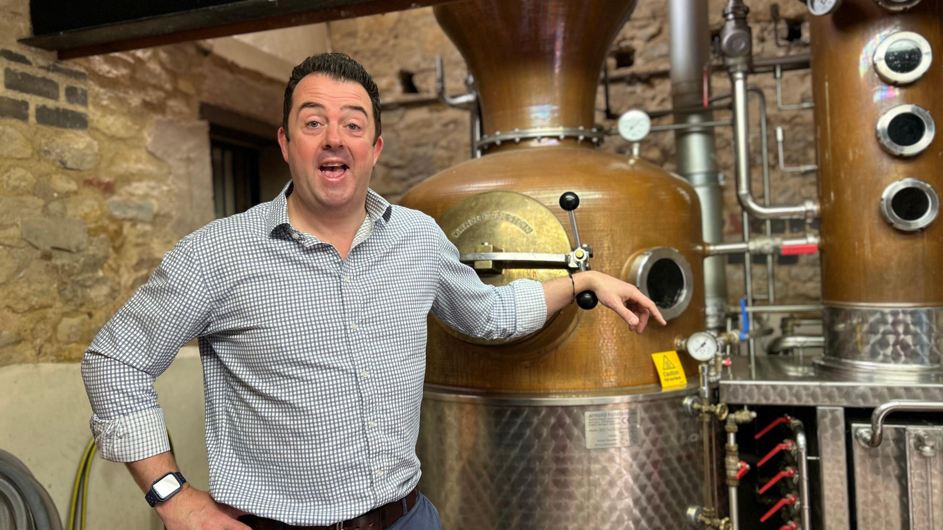 A man in a white and black chequered shirt and jeans stands next to a large bronze gin distiller. 