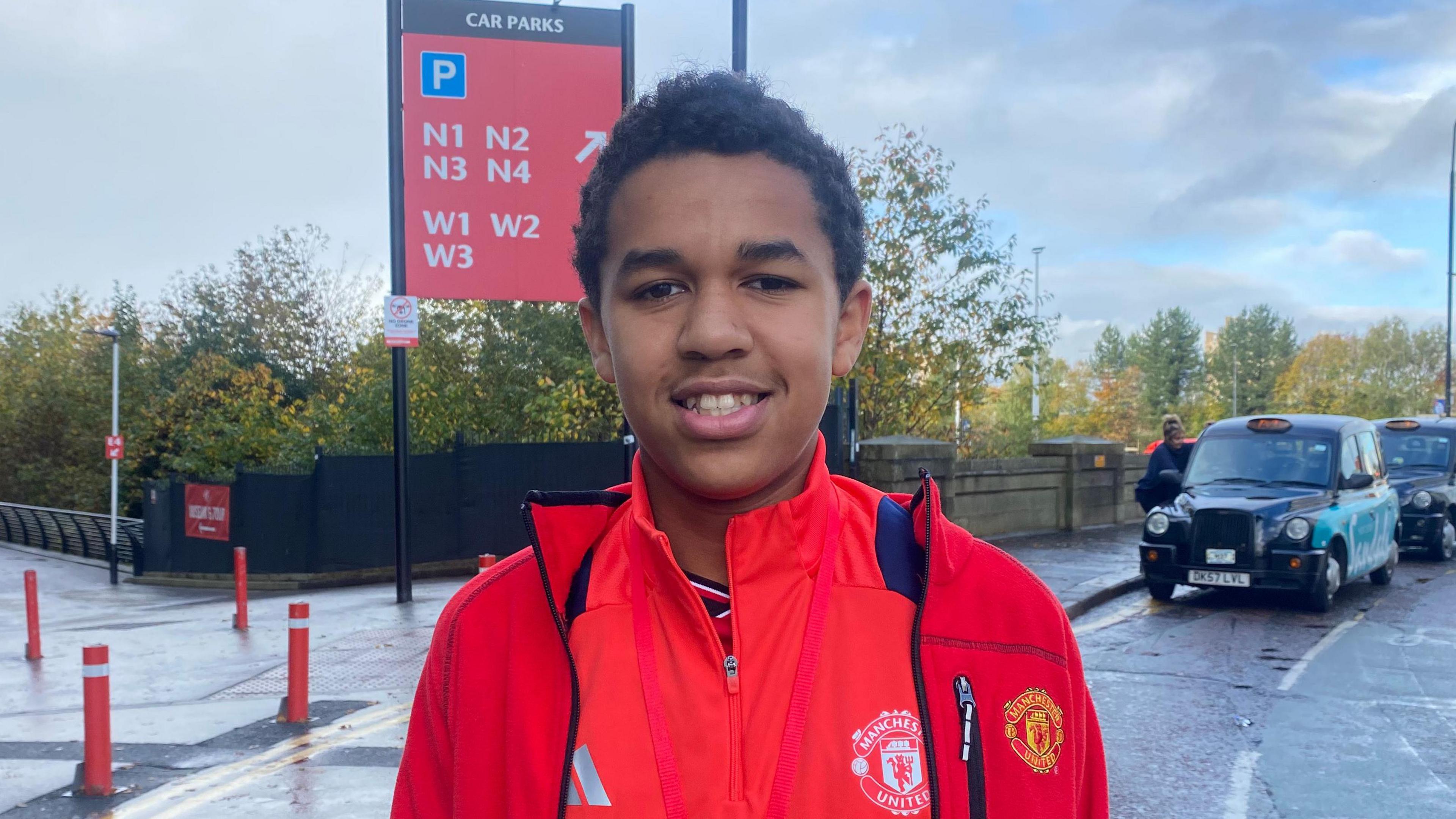 Alexander, a 12-year-old Manchester United fan, wearing a red club jacket, smiling for the camera outside a roadway. 