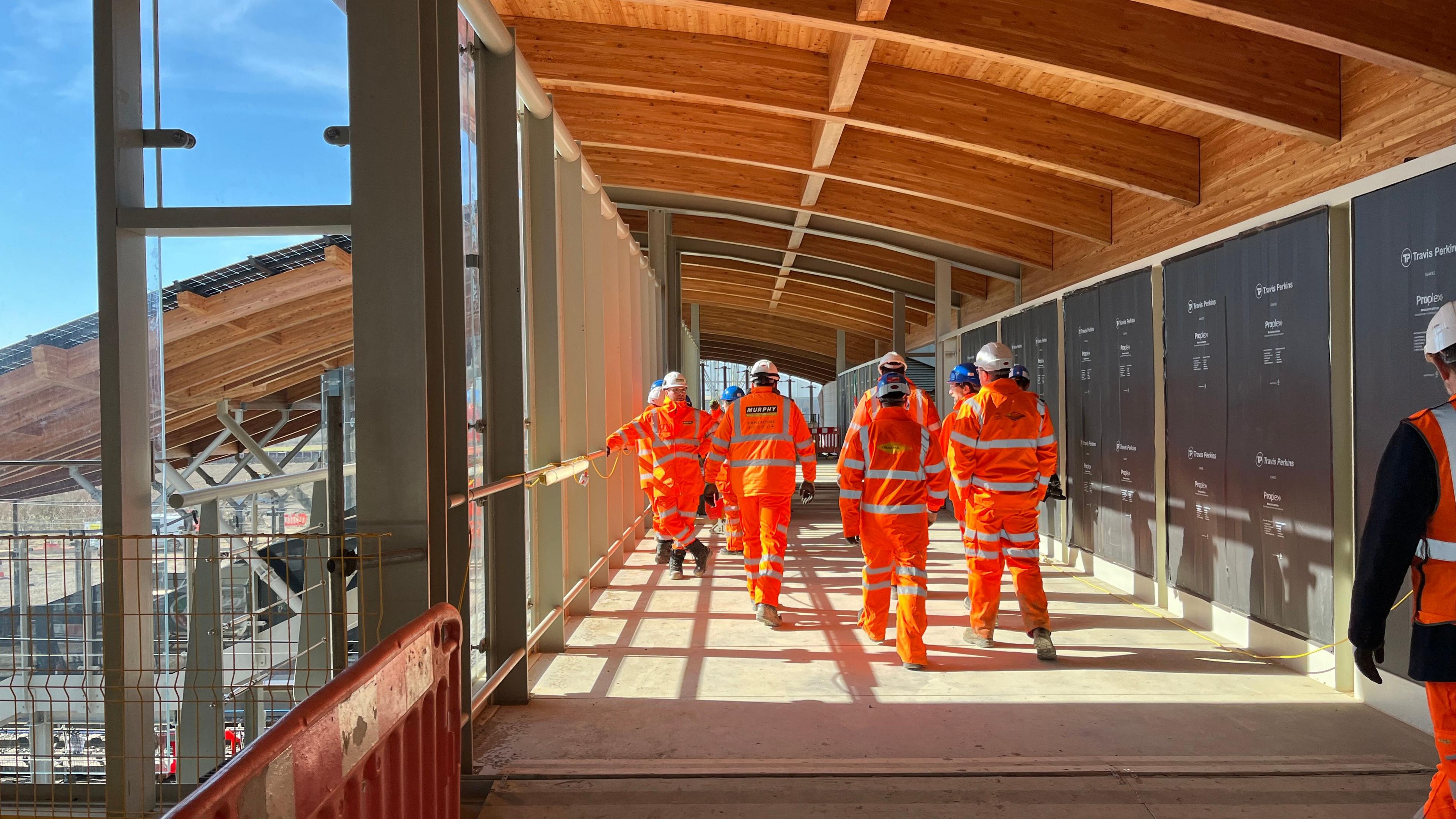 Construction workers wearing orange hi-vis and hard hats are walking on one of the storeys of the station, there is a safety barrier in the foreground.