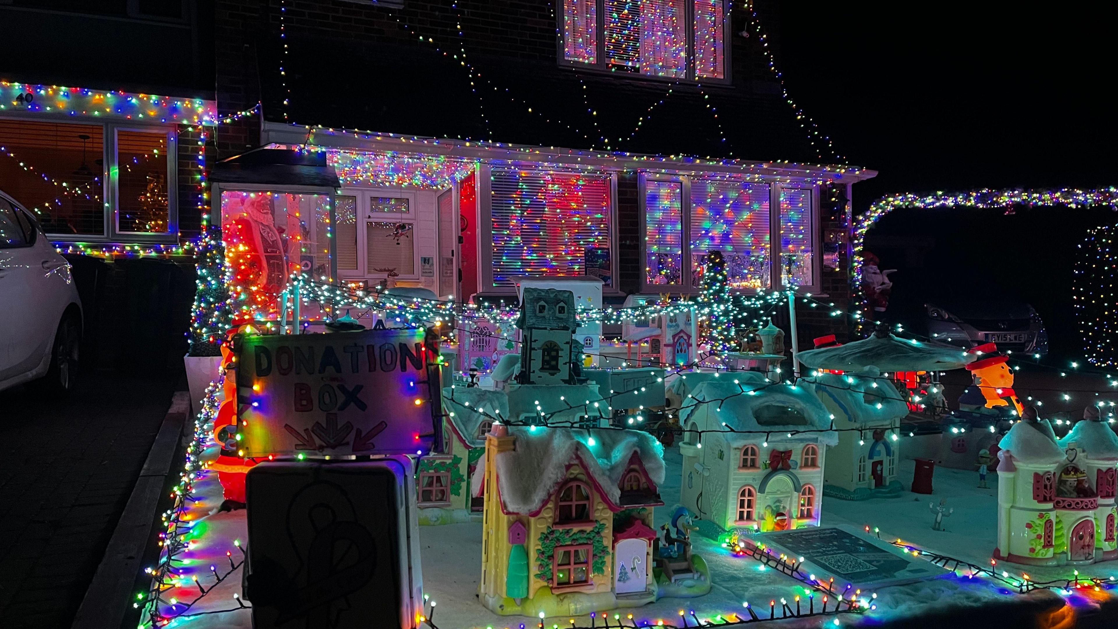 A house is festooned in thousands of Christmas lights, with a display in the foreground that includes a model village covered in fairy lights.