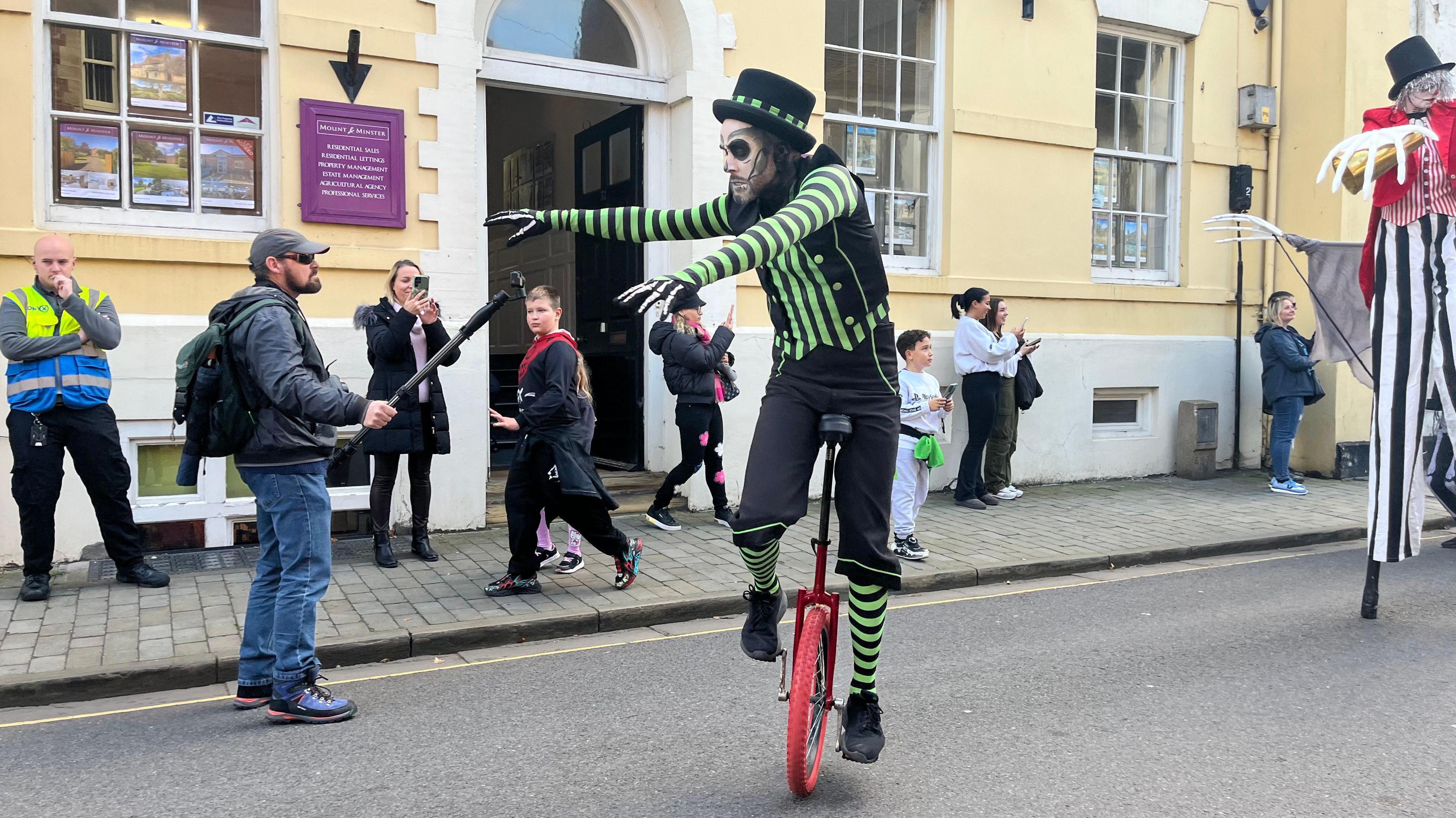 A man riding a unicycle wearing a green and black skeleton outfit in the streets of Lincoln. Members of the public are watching and taking photos. He has skeleton face paint on.