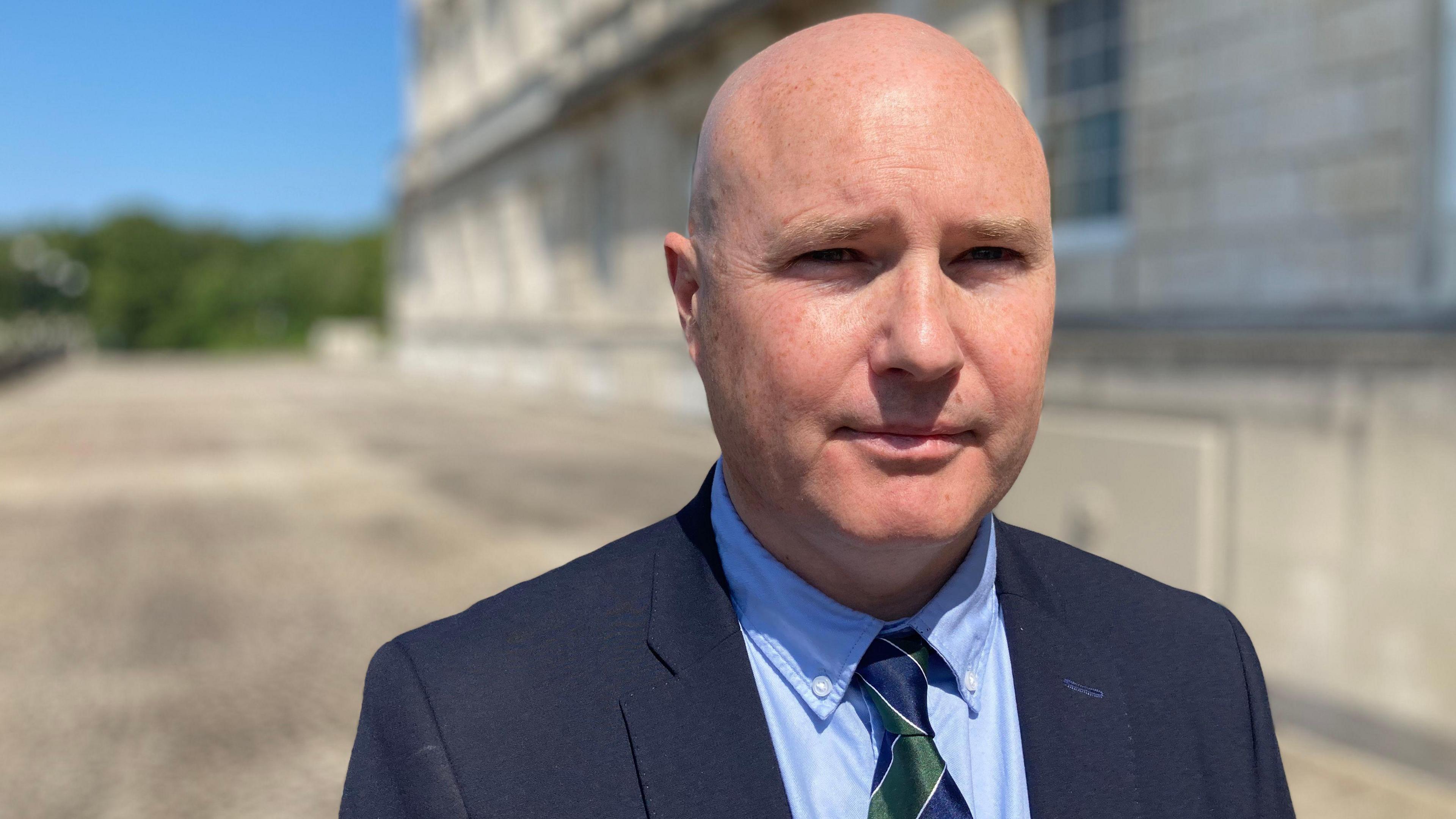 A bald man is wearing a navy suit with a light blue shirt and a green and blue striped tie. He is bald and has blue eyes. He is looking towards the camera with a serious expression. He is standing before the Stormont assembly building on a blue sunny day.