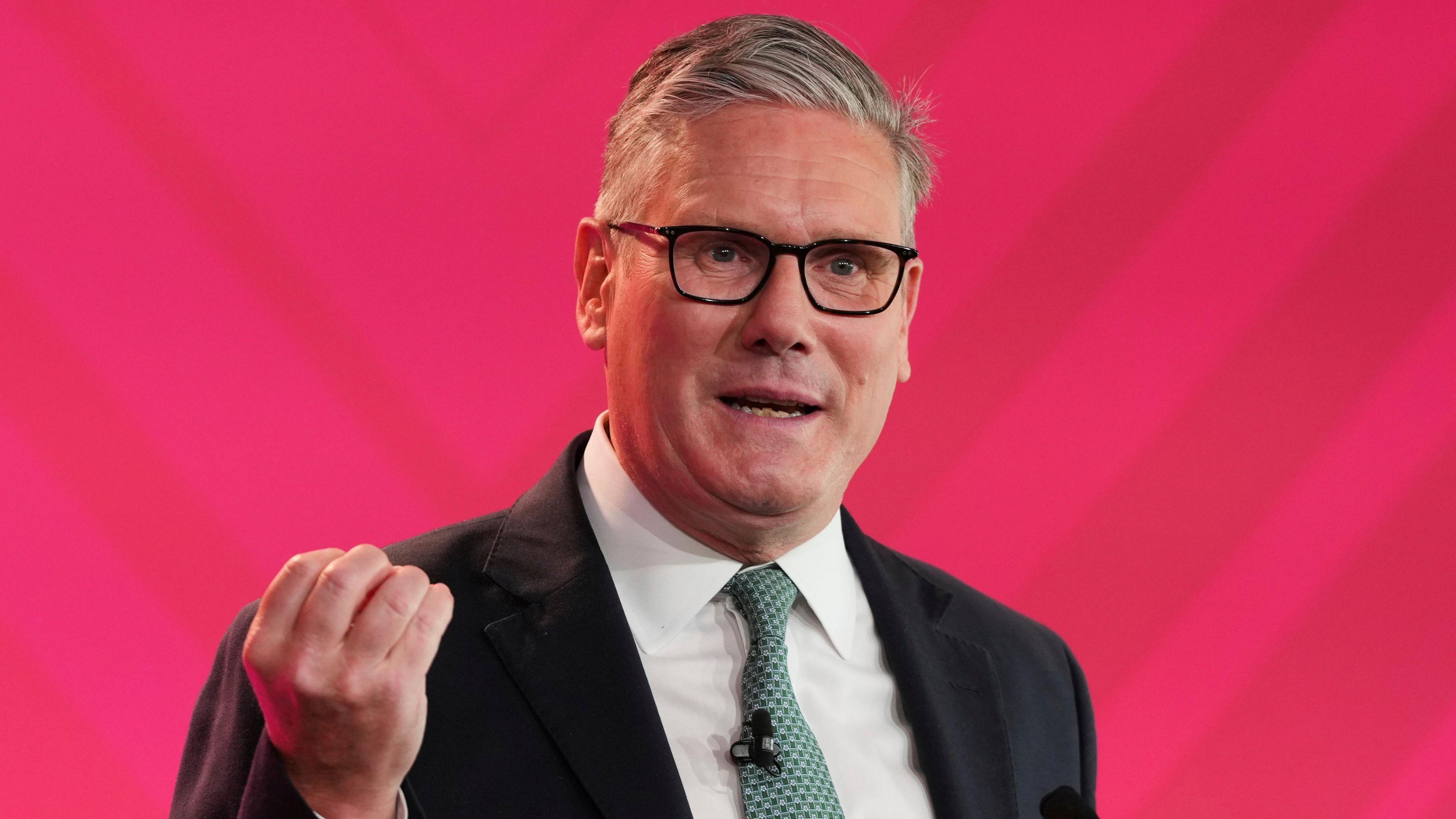 Sir Keir Starmer holds up his right hand while making a speech in front of a pinkish-red background. He wears a pale green tie.