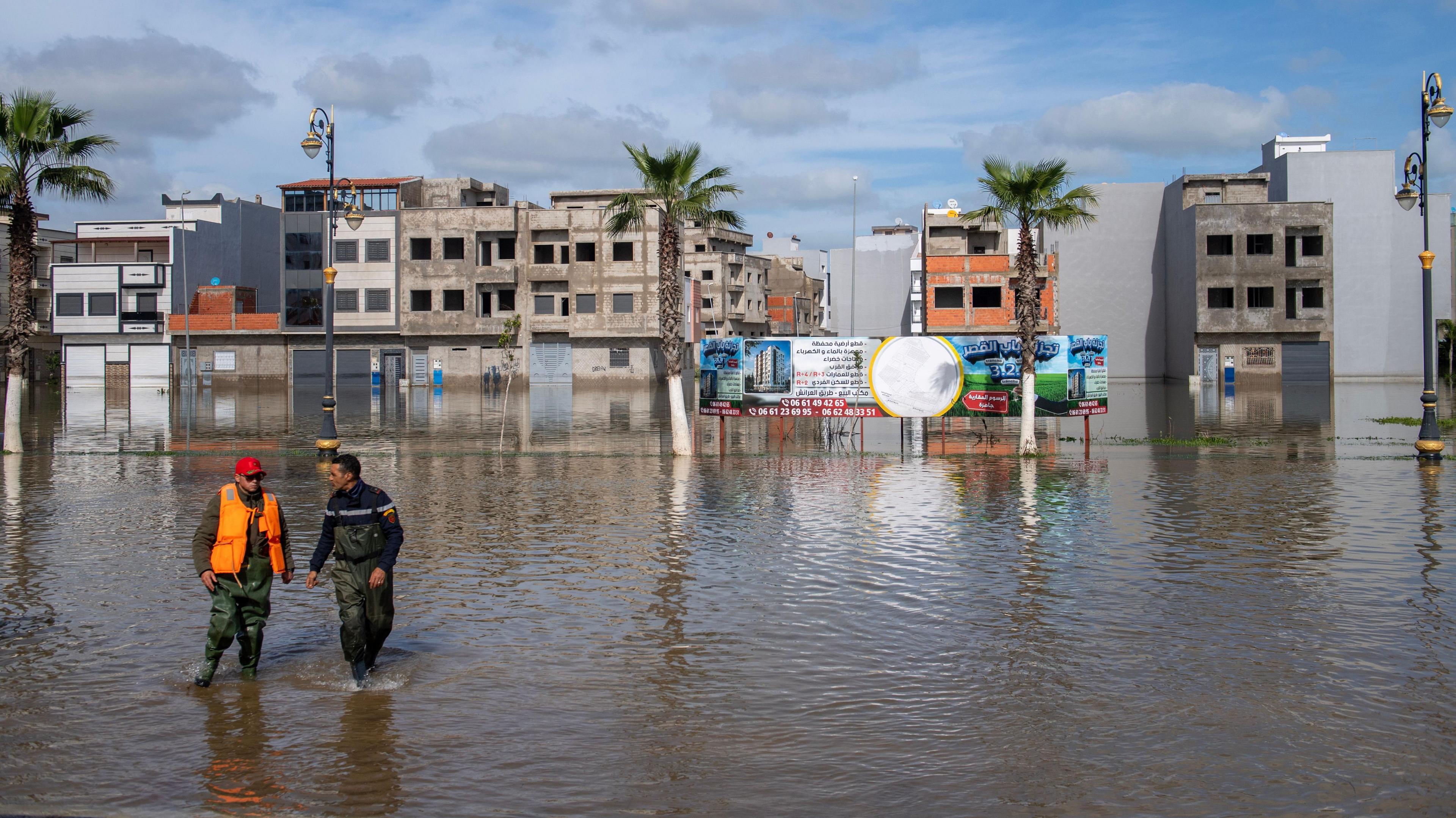Buildings and palm trees. Two men wade in a flooded street.
