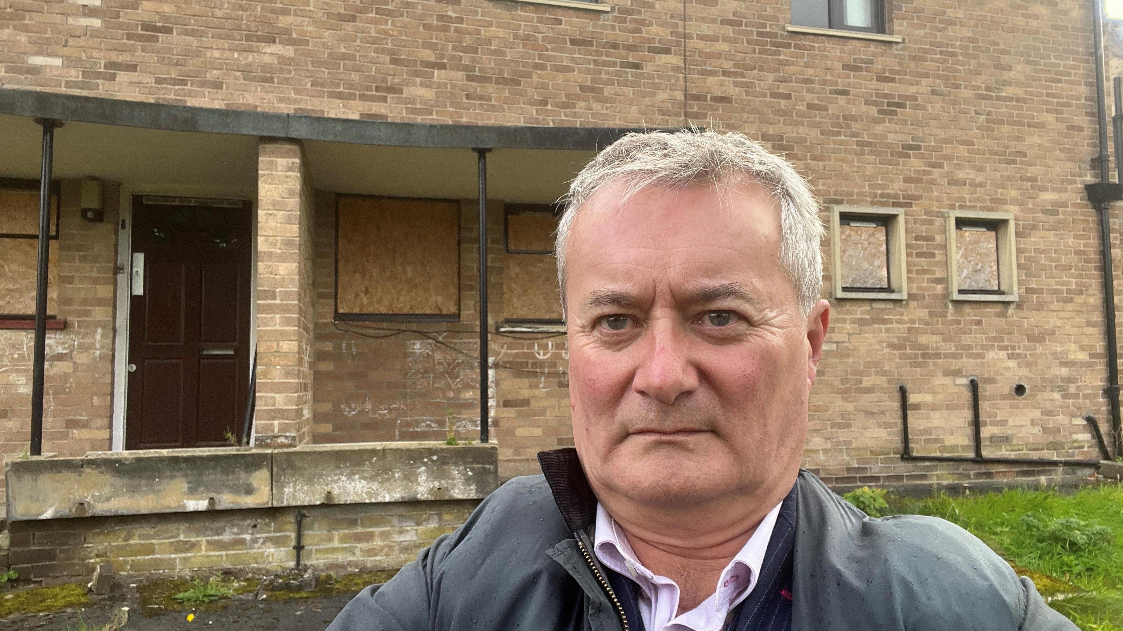 Head and shoulder shot of a man with a serious expression, short greying hair and a grey anorak standing in front of a boarded up municipal-looking brick building.